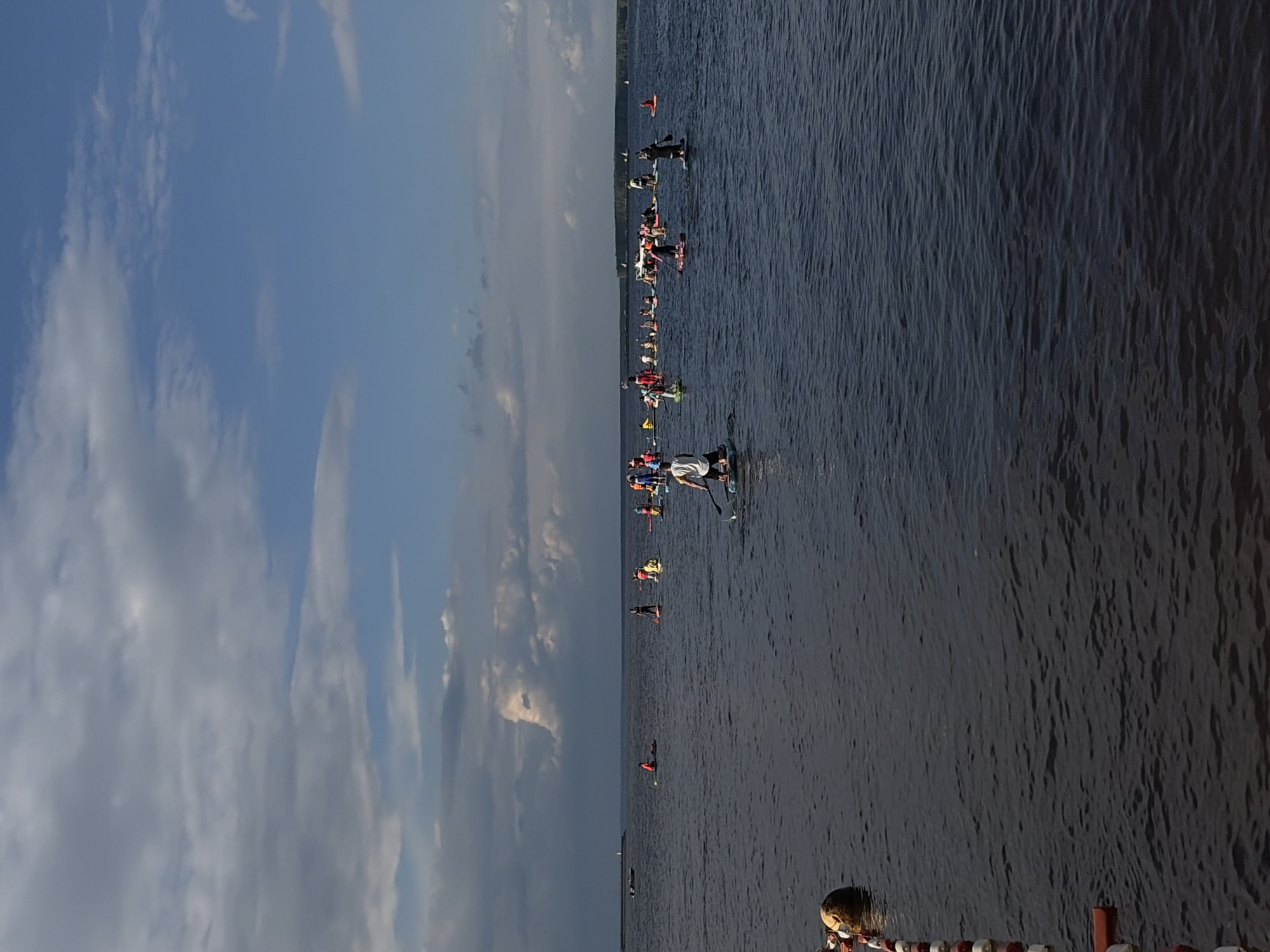 The image depicts a wide shot of a body of water with a shoreline in the distance, under a sky with scattered clouds. The scene is dominated by the dark, rippled surface of the water, reflecting the muted light of the sky. A large group of people on paddleboards are spread across the water, participating in what appears to be a paddleboard race or event.
The individuals are scattered, with some grouped together and others further apart. They are all standing on paddleboards, holding paddles, and some are wearing life vests in bright colors like orange and yellow, suggesting safety precautions for the water activity. The people are diverse in their attire, with a mix of colorful and neutral clothing visible.
In the foreground, on the right side of the image, part of a wooden structure with red and white stripes and a round, brown object, possibly a buoy, are visible, suggesting the edge of a dock or event area. Further in the distance, along the horizon, a thin line of trees and buildings indicates the shoreline.
The sky transitions from a bright blue on the left, with wispy white clouds, to darker, more overcast conditions towards the right. The lighting suggests it is likely late afternoon or early evening, given the soft light and the shadows cast on the water. The overall atmosphere appears to be one of active participation and potentially friendly competition. No visible text is present in the image.