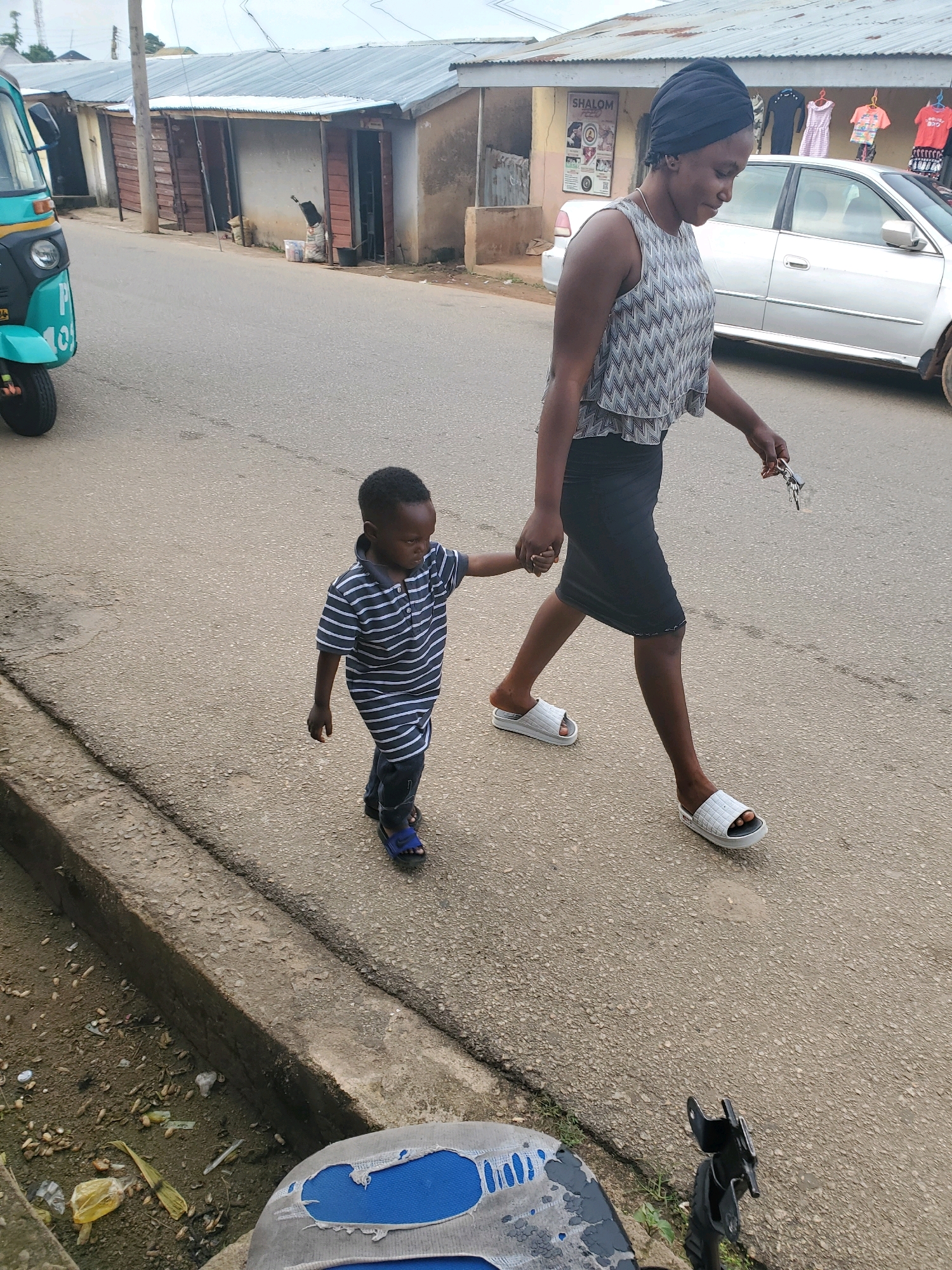 A woman wearing a black headwrap, a patterned sleeveless top, and a black skirt walks on a paved street in Jos, Nigeria, holding the hand of a young boy. The boy, wearing a striped t-shirt and dark pants, walks beside her, also in the street. In the background, a silver car is parked in front of a row of shops with corrugated metal roofs. A blue and yellow auto-rickshaw is visible on the left side of the frame. The street is lined with a concrete gutter.