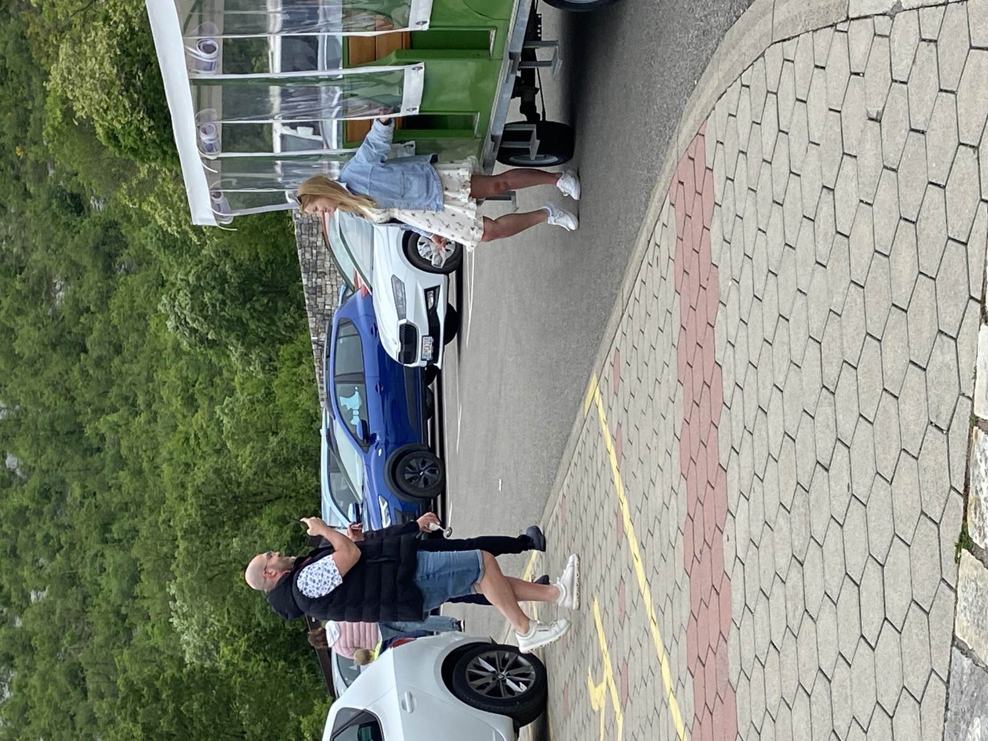 The image captures a scene in Lipa Dobrska, Montenegro. A woman with blonde hair, wearing a white dress with floral patterns and a light blue denim jacket, stands near the side of a green and white open-air tour vehicle with a clear plastic roof. She appears to be either entering or exiting the vehicle. Adjacent to the vehicle are parked cars, including a white Skoda and a blue car.
In the foreground, a man with a bald head and glasses is standing and gesturing with his right hand, while his left hand holds an object. He is wearing a black vest over a white shirt with a floral pattern, along with blue shorts and white sneakers. Another person, partially visible, is wearing a pink top.
The scene is set in a parking area with a paved surface made of gray hexagonal blocks with a border of red blocks and a yellow line marking the edge. The background features dense green trees, indicating a natural landscape surrounding the parking area.