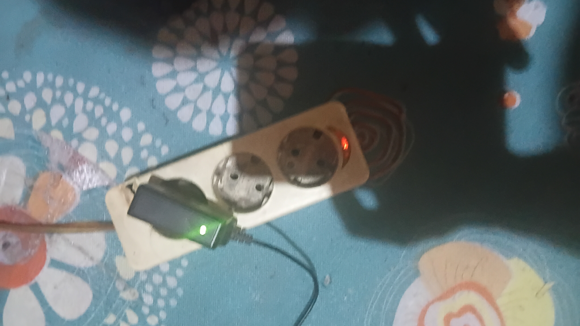 The image shows a power strip with two outlets on a patterned surface. A black power cord is plugged into the power strip, leading to a green electronic device with a lit indicator light. One of the outlets on the power strip has a faint red light emanating from it. The background has a teal color with a white floral pattern and some abstract circular designs in orange and brown. Shadows are cast on the surface, suggesting artificial lighting. The location is identified as Prabumulih, Indonesia, though no specific landmarks or contextual clues within the image support this geographical assignment beyond the provided metadata.