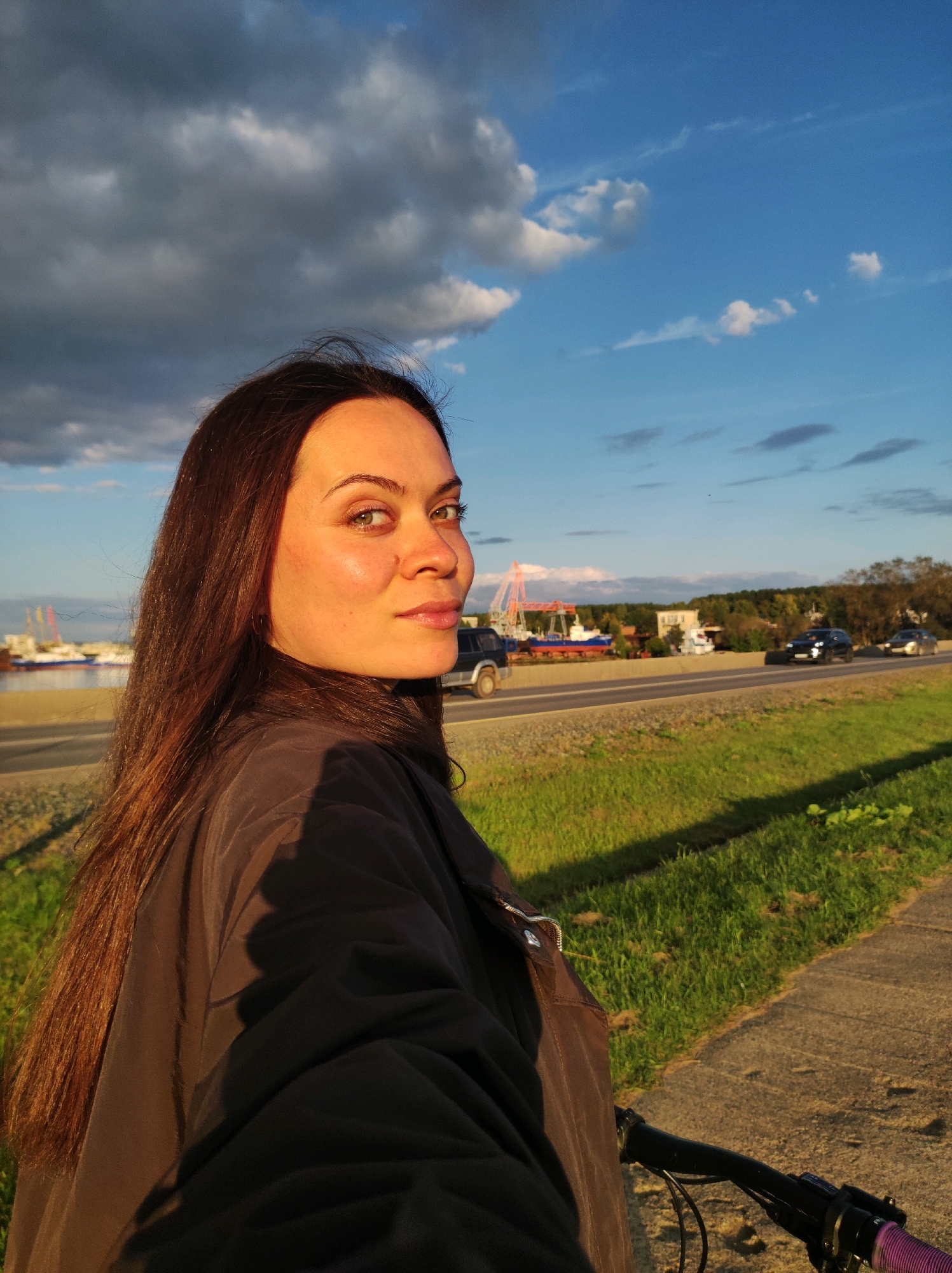 An outdoor, eye-level, medium shot captures a woman from the shoulders up, looking over her right shoulder at the camera. Her long, dark brown hair is loose and falls past her shoulders. She wears a dark brown jacket. The woman is positioned on the left side of the frame. The background features a paved pathway on the right, with a grassy embankment next to it. Beyond the embankment is a highway with cars driving on it. In the distance, there are industrial structures, including cranes and ships near a body of water, possibly the Kama River, indicating the location is in Chaikovsky, Russia. The sky is a mix of blue with scattered clouds, illuminated by the warm glow of late afternoon or early evening sun. The scene appears to be a cyclist's perspective, with the handlebars of a bicycle visible in the lower right corner of the frame.