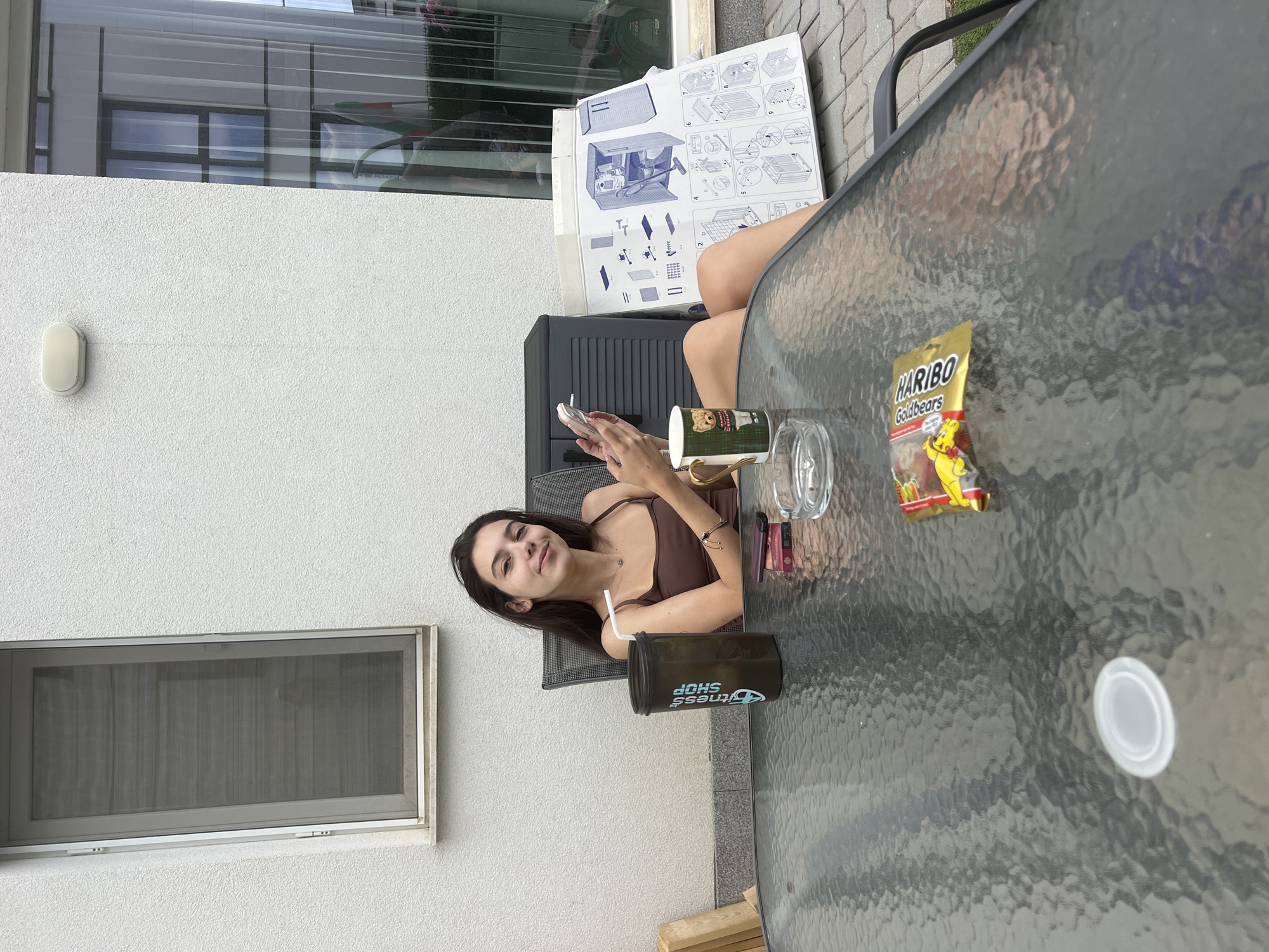 A young woman with dark hair is sitting at a glass table outdoors, holding a cigarette in her right hand and a cell phone in her left. She is wearing a brown tank top. To her left on the table is a black protein shaker with text that says "FITNESS SHOP". Next to the shaker is a mug with a teddy bear design. Further to the right, there is an ashtray with a cigarette butt, a pack of cigarettes, and a bag of Haribo Goldbears. In the background, there is a grey storage unit and a large piece of cardboard with assembly instructions for a storage unit. The scene takes place on a patio next to a building with white textured walls and a window.