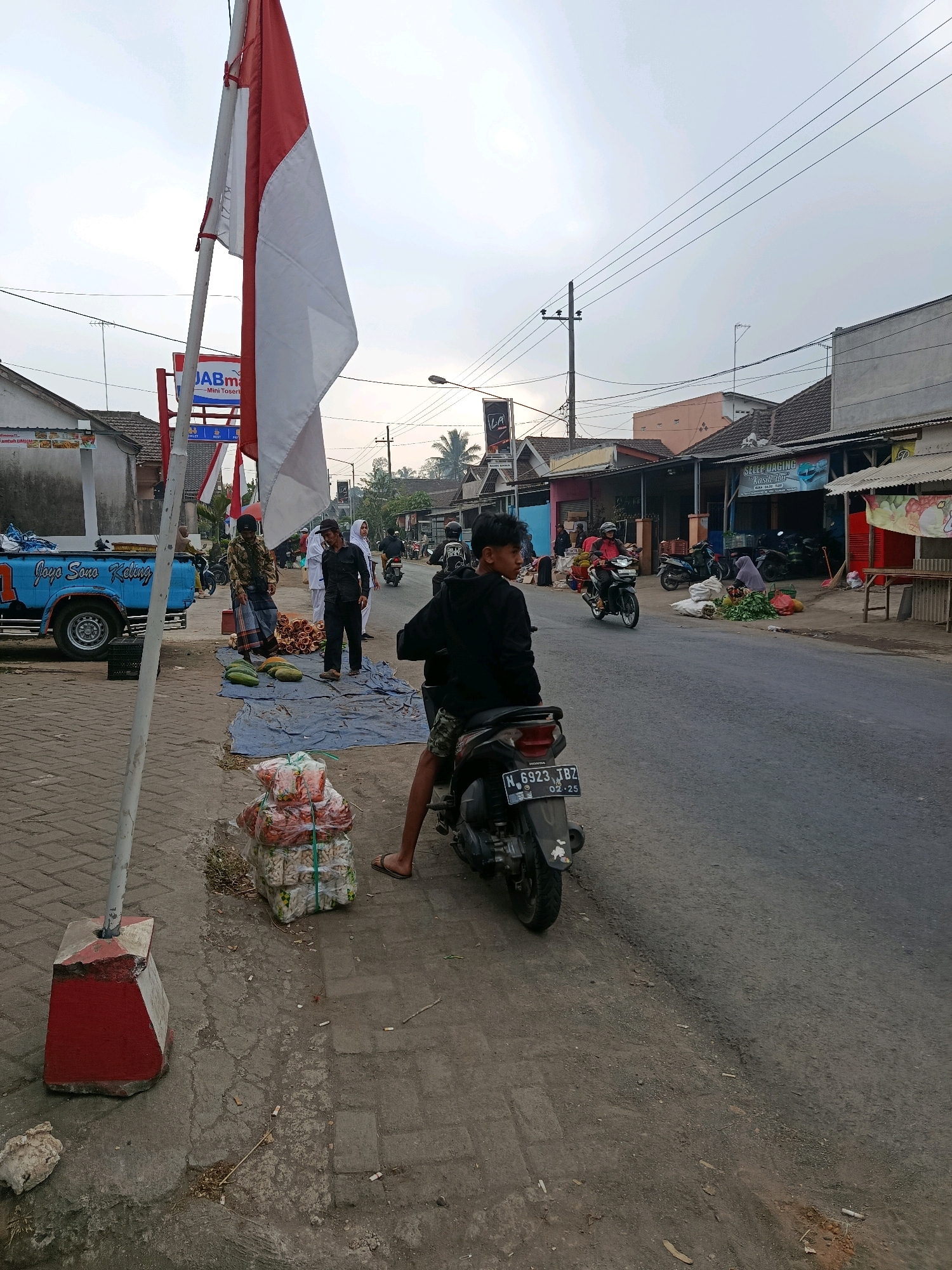 This image depicts a street scene in Tajinan, Indonesia. In the foreground, a young man on a motorcycle faces away from the camera, with the rear of his vehicle visible. To his left, a blue pickup truck is parked on a brick-paved sidewalk. A large Indonesian flag, red and white, is prominently displayed on a pole to the left of the frame. A group of people are gathered near the truck, with some standing and others tending to goods laid out on a blue tarp on the ground. These goods appear to be produce, including what look like cucumbers and other vegetables. Piles of bagged items, likely groceries or merchandise, are stacked on the sidewalk near the flag pole.
The street itself is paved and appears to be a busy thoroughfare, with several other motorcycles and a few pedestrians visible in the background. Shops and buildings line the right side of the street, some with awnings and signage. Power lines are strung across the sky. The overall atmosphere suggests a typical market day or roadside commerce in a residential or commercial area. The sky is overcast, indicating potentially cloudy or hazy weather conditions.