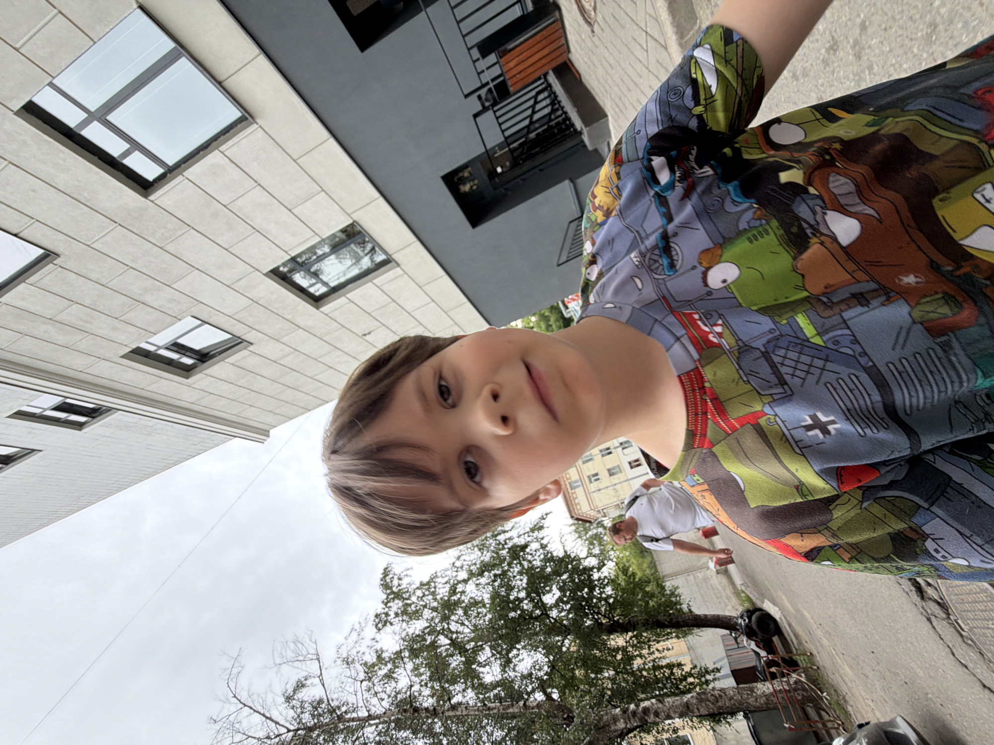 This is a selfie of a young boy with light brown hair and brown eyes, looking directly at the camera. He is wearing a colorful t-shirt with cartoon characters printed on it. The photo is taken from a low angle, with the boy's face prominently featured at the bottom of the frame.
The background shows a street scene with buildings and trees. To the left is a modern building with large, rectangular windows. To the right, there is another building with balconies and orange accents. A man wearing a white shirt and a backpack is walking on the sidewalk in the middle ground. The sky is overcast, suggesting a cloudy day. The overall setting appears to be an urban environment.
The boy's expression is neutral, and the lighting suggests it might be daytime, possibly late morning or early afternoon given the overcast sky. No text is visible in the image. The composition and low angle of the selfie, along with the surrounding architecture, hint at a possibly residential or commercial area in Moscow, Russia.