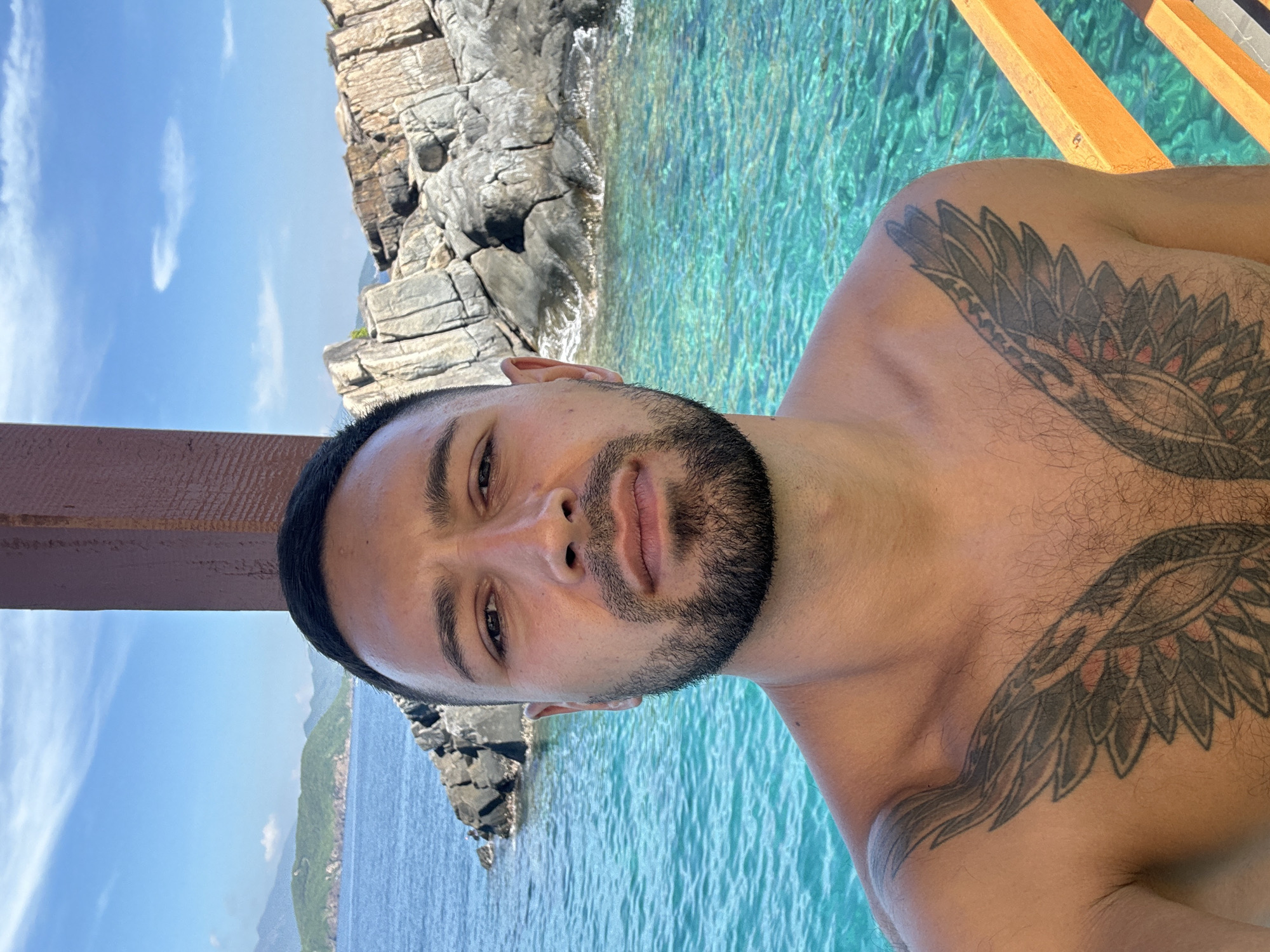 A close-up, chest-up selfie is taken by a man with a beard and dark hair, standing on a wooden deck overlooking a body of turquoise water. His chest displays a large, black wing tattoo. In the background, rocky islands with lush green vegetation rise from the water under a bright blue sky with scattered white clouds. The man appears to be in the city of Nha Trang, Ward, Vietnam.