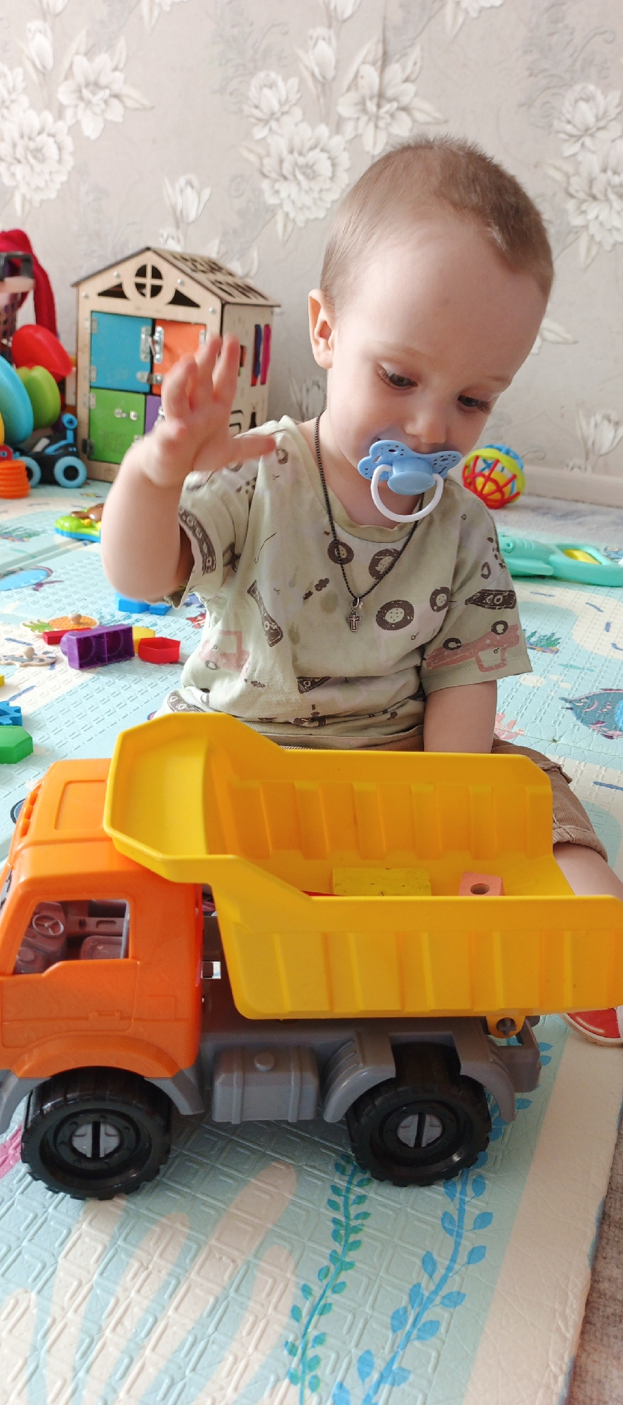 A young child, likely a toddler, is sitting on a colorful play mat indoors, engaged in play. The child has light brown hair and is wearing a light green t-shirt patterned with what appear to be vehicles or abstract shapes. A blue pacifier is in the child's mouth, and a black necklace with a small silver cross pendant hangs around their neck. Their right hand is raised and slightly open, seemingly in mid-motion. The child's gaze is directed downwards, focused on the toys.
In the foreground, a large orange and yellow toy dump truck is prominently featured. The truck has an orange cab and a bright yellow tipping bed, which contains a few small, colorful building blocks, including a yellow rectangular one and a pink cylindrical one. Various other toys are scattered around the child on the play mat, including multiple colorful building blocks (purple, red, green, blue) to the left, and a multi-colored, open-weave ball further back on the right. Behind the child on the left, a wooden "busy board" house with different colored doors and latches is visible. The play mat itself has a light blue and white pattern with leaf-like designs. The background features light-colored wallpaper adorned with a floral pattern of white flowers. The scene depicts a child engrossed in playtime within a home setting in Chaikovsky, Russia. There is no visible text.