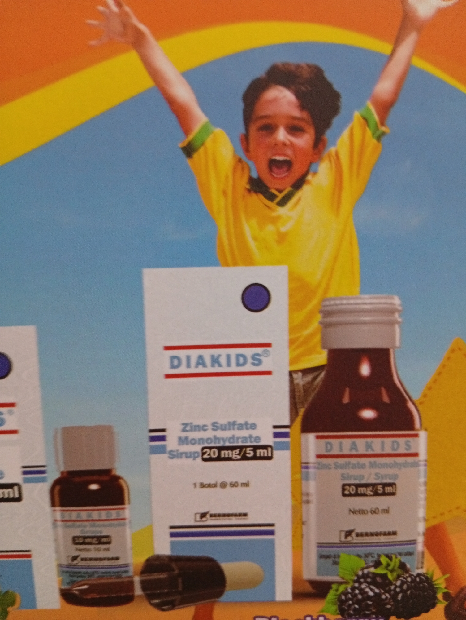 The image shows a product advertisement for "DIAKIDS" syrup, featuring a young boy in a yellow shirt with his arms raised in excitement. The product packaging is visible, with bottles and a box detailing "Zinc Sulfate Monohydrate Syrup." The background is a vibrant blue sky with yellow and orange abstract shapes. The advertisement appears to be for a product sold in Indonesia, potentially in Tlogomulyo. Several bottles of the syrup are displayed, along with a close-up of blackberries, suggesting a fruit flavor. The overall impression is one of a cheerful and healthy product targeting children.