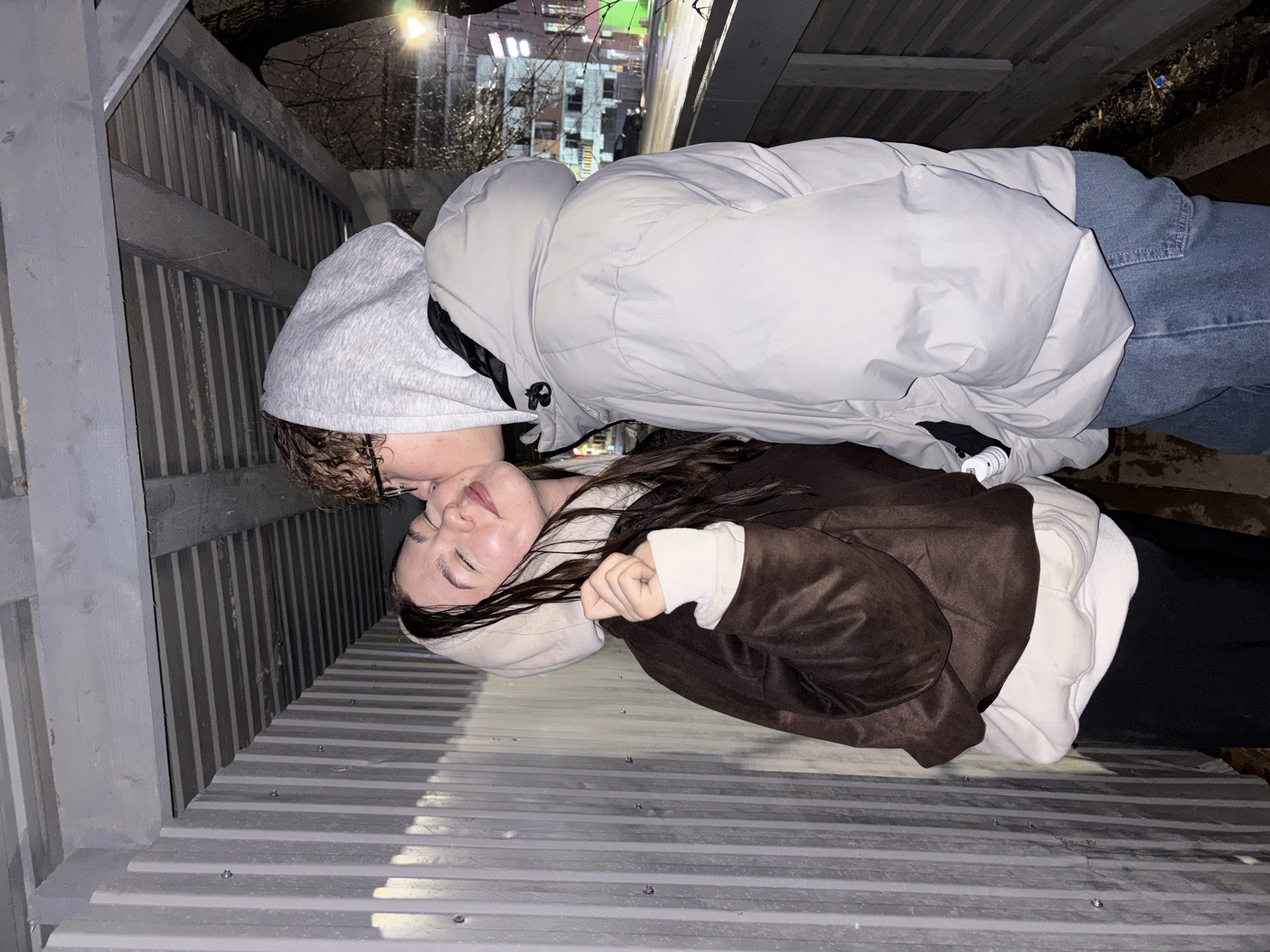 A couple is lying on a corrugated metal surface under a structure. One person, presumably male, is wearing a gray hooded sweatshirt and a light gray puffer jacket with jeans. The other person, presumably female, is wearing a white sweatshirt under a brown jacket and black pants. The couple appears to be kissing. The location is under an elevated structure, possibly a pedestrian walkway or bridge. In the background is an urban scene with buildings and streetlights, possibly in the city of Perm, Russia. The image has been rotated so the couple is sideways. The lighting suggests it is nighttime.
