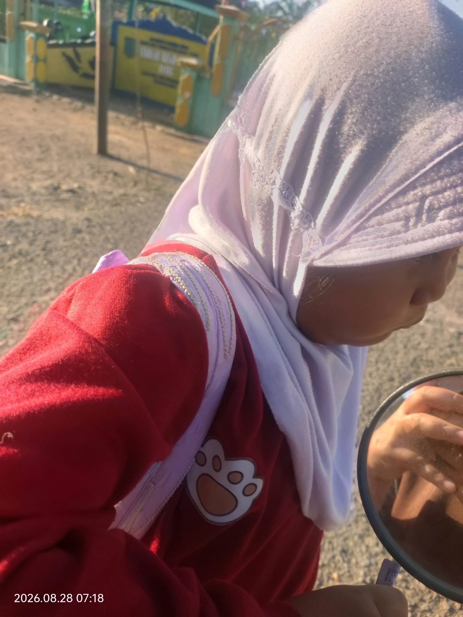 An outdoor, close-up, low-angle, eye-level shot shows a child wearing a red top with a white, paw-print design and a white hijab. The child is looking into a circular mirror, with their right hand visible in the reflection. The background is blurred but suggests a rural or outdoor setting with a dirt ground and a structure with yellow and green panels in the distance. A timestamp in the bottom left corner reads "2026.08.28 07:18".