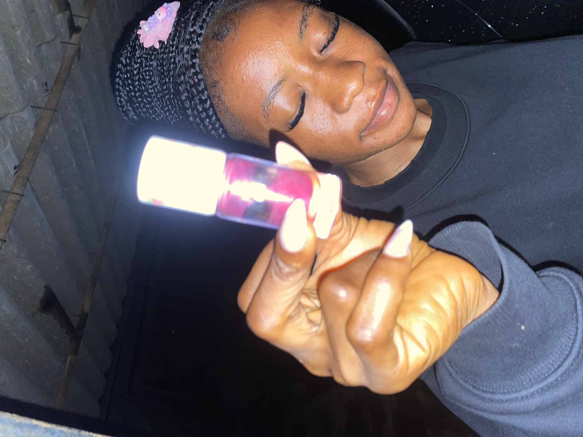 A close-up shot focuses on the face and hand of a young Black woman, who is lying down with her eyes closed and a slight smile. Her hair is styled in braids, adorned with a small pink accessory. She is holding a small, cylindrical object that is illuminated from within, casting a bright light. The object appears to contain a pink liquid.
The woman is wearing a black crewneck sweatshirt. Her fingernails are painted white, and the lighting highlights the texture of her skin. The background is dark and appears to be an outdoor, possibly dimly lit environment. On the left side of the image, there is a textured, possibly concrete or brick wall, with some horizontal wooden beams or supports visible. The overall impression is one of a relaxed or introspective moment, perhaps at night, given the darkness and the illuminated object. No visible text is present in the image. The context suggests this is in Jos, Nigeria.