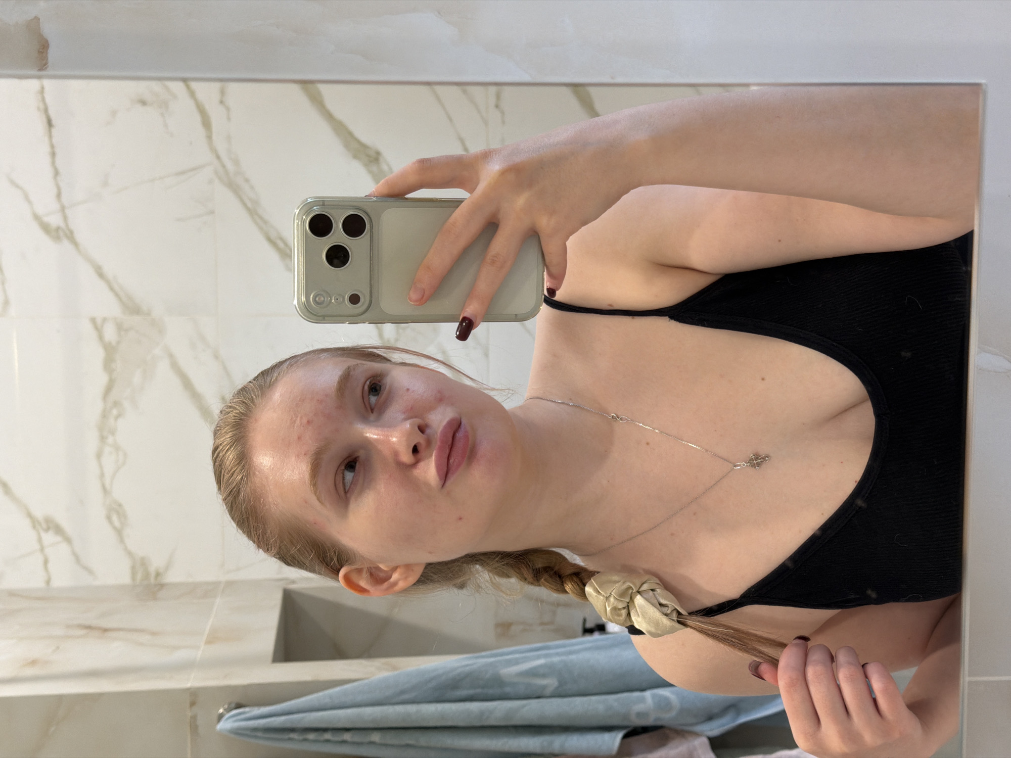 A fair-skinned woman is taking a selfie in a bathroom. She is wearing a black top and a silver necklace with a cross pendant. Her hair is blonde and braided, held together with a tan scrunchie. She is holding a grey smartphone. The bathroom features marble-patterned walls and a blue towel is visible. This does not appear to be in the city of Rossiyskiy, Russia.