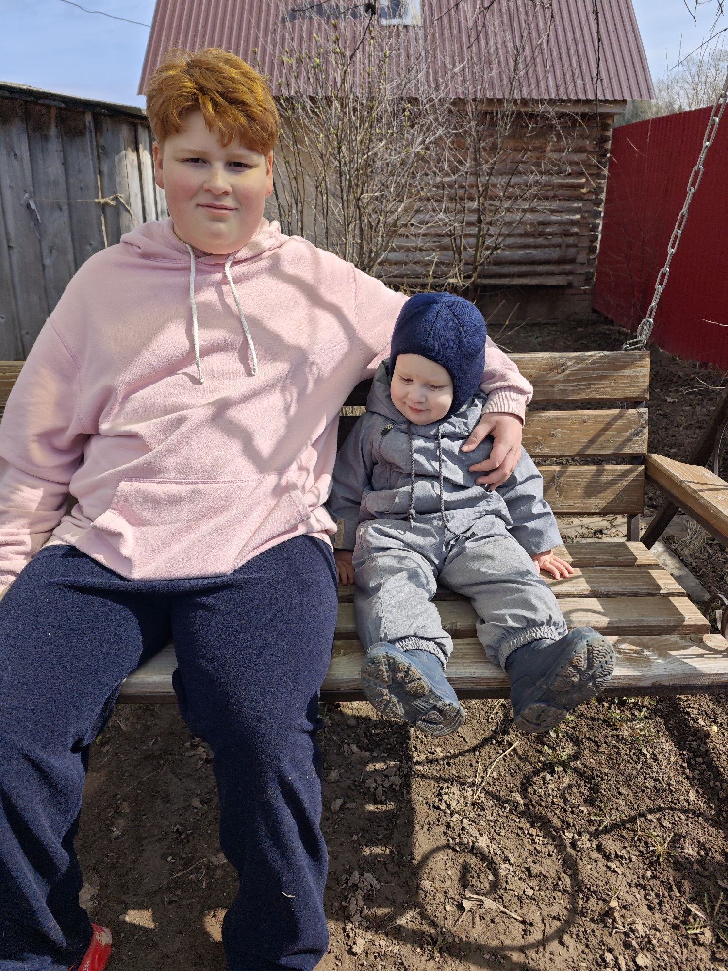 Two children, one older than the other, are sitting on a wooden swing. The older child, who appears to be a boy, has red hair and is wearing a pink hooded sweatshirt and dark blue pants. His arm is around the smaller child, who is wearing a gray winter suit with a dark blue knit hat. Both children are looking down. The swing is suspended by chains. The ground beneath the swing is bare soil. In the background is a wooden fence, a log cabin, and a red metal fence. The location is likely in a rural or suburban area near the city of Фоки, Russia.