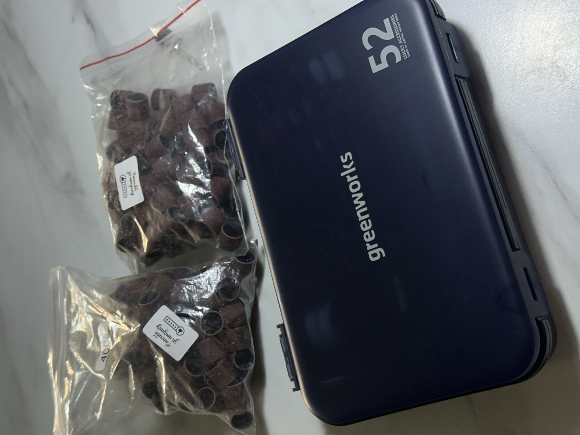 The image displays a dark blue accessory case and two clear plastic bags filled with sanding bands, all placed on a light-colored, possibly marble surface.
The rectangular, dark blue case, likely made of plastic, features the brand name "greenworks" printed vertically in white on its side. At the top right, the number "52" is prominently displayed in white, accompanied by smaller text that reads "LUCKY ACCESSORIES" and "SMART AV ELECTRIC ROTARY TOOLS," indicating its purpose for storing rotary tool accessories.
Next to the case are two transparent, resealable plastic bags, each containing a quantity of small, brown, cylindrical sanding bands or sleeves. These are commonly used with rotary tools for sanding or grinding. Both bags have white rectangular stickers. The sticker on the upper bag partially shows "Спасибо за покупку" (Thank you for your purchase) and a five-star rating graphic with "faniform of". The sticker on the lower bag clearly displays "400" which typically denotes a grit size for abrasives, and also contains the partially visible "Спасибо за покупку" and five-star rating.
The overall scene suggests an indoor setting with bright, even lighting. There are no people present, and the image focuses solely on these items. The objects appear to be new or well-maintained, implying readiness for use or storage.