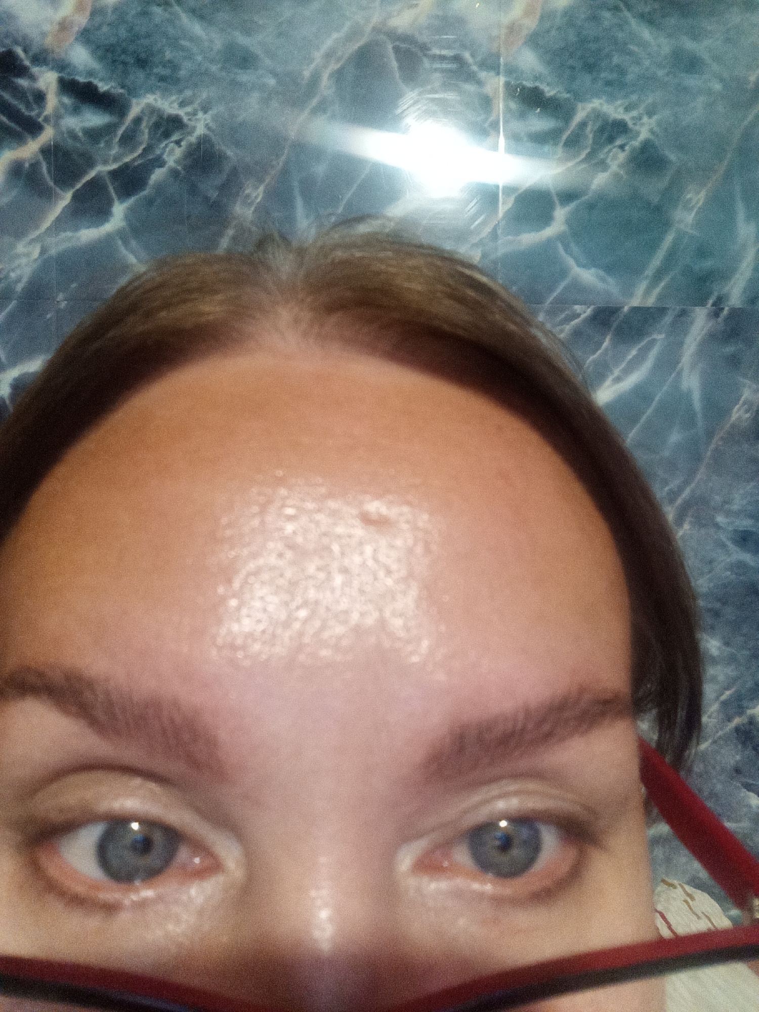 A close-up, eye-level medium shot captures the top half of a person's face, focusing on their forehead, eyebrows, and eyes. The individual has fair skin and light brown hair that is parted in the middle and pulled back. Their eyebrows are well-defined and dark. The eyes are open and appear to be blue-gray, with visible reflections in the pupils. The person is wearing red-framed glasses, which are partially visible at the bottom of the frame. The background is a marbled pattern of blue and white. No specific location in Novopokrovka, Russia, can be identified from the image.