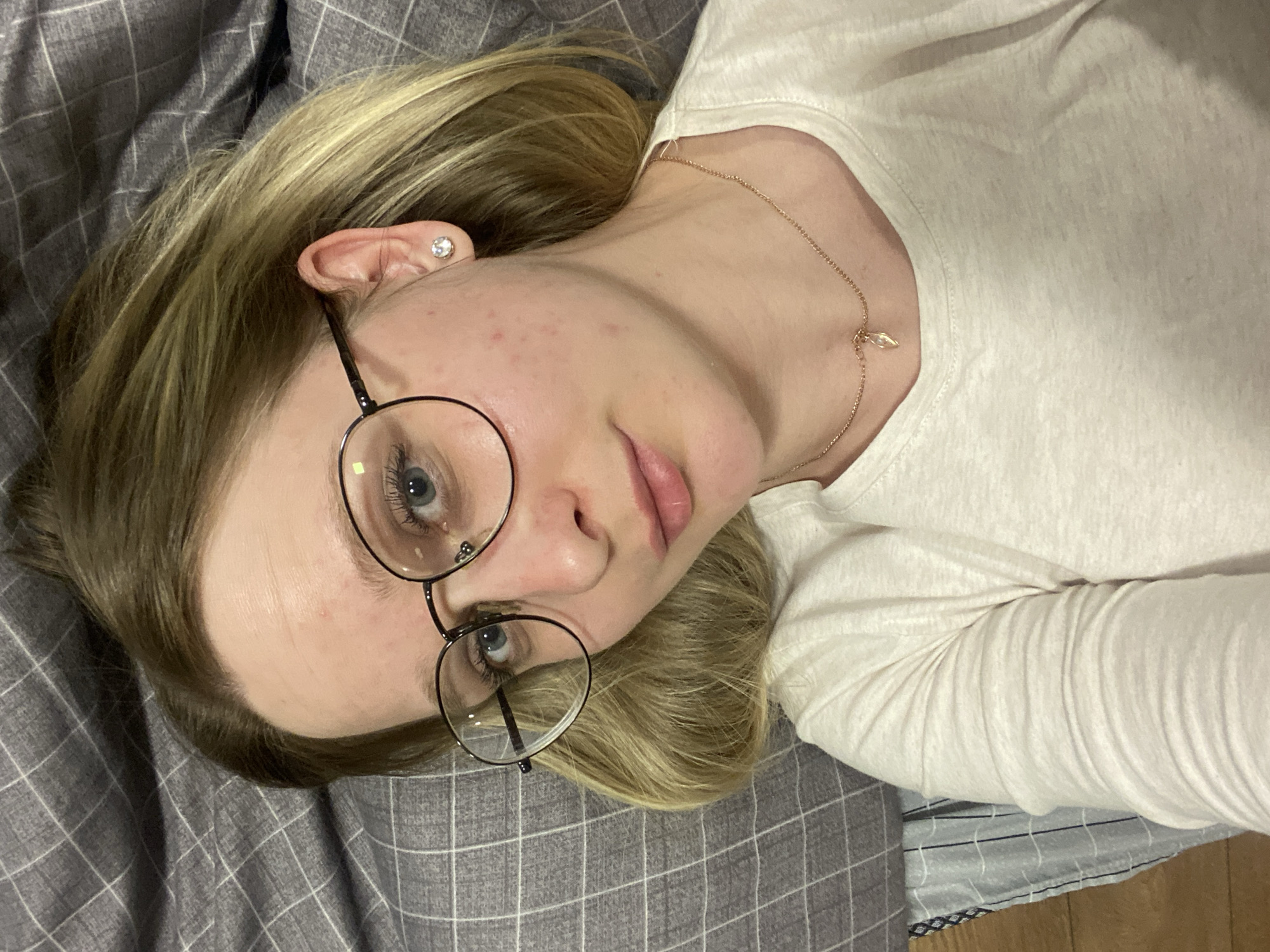 A young woman with blonde hair and blue eyes is lying on a gray checkered bedsheet. She is wearing a white long-sleeved shirt, round black-rimmed glasses, and a delicate gold necklace with a small pendant. She has a small earring in her left ear. Her skin shows some minor blemishes. The location is not identifiable as Izhevsk, Russia.