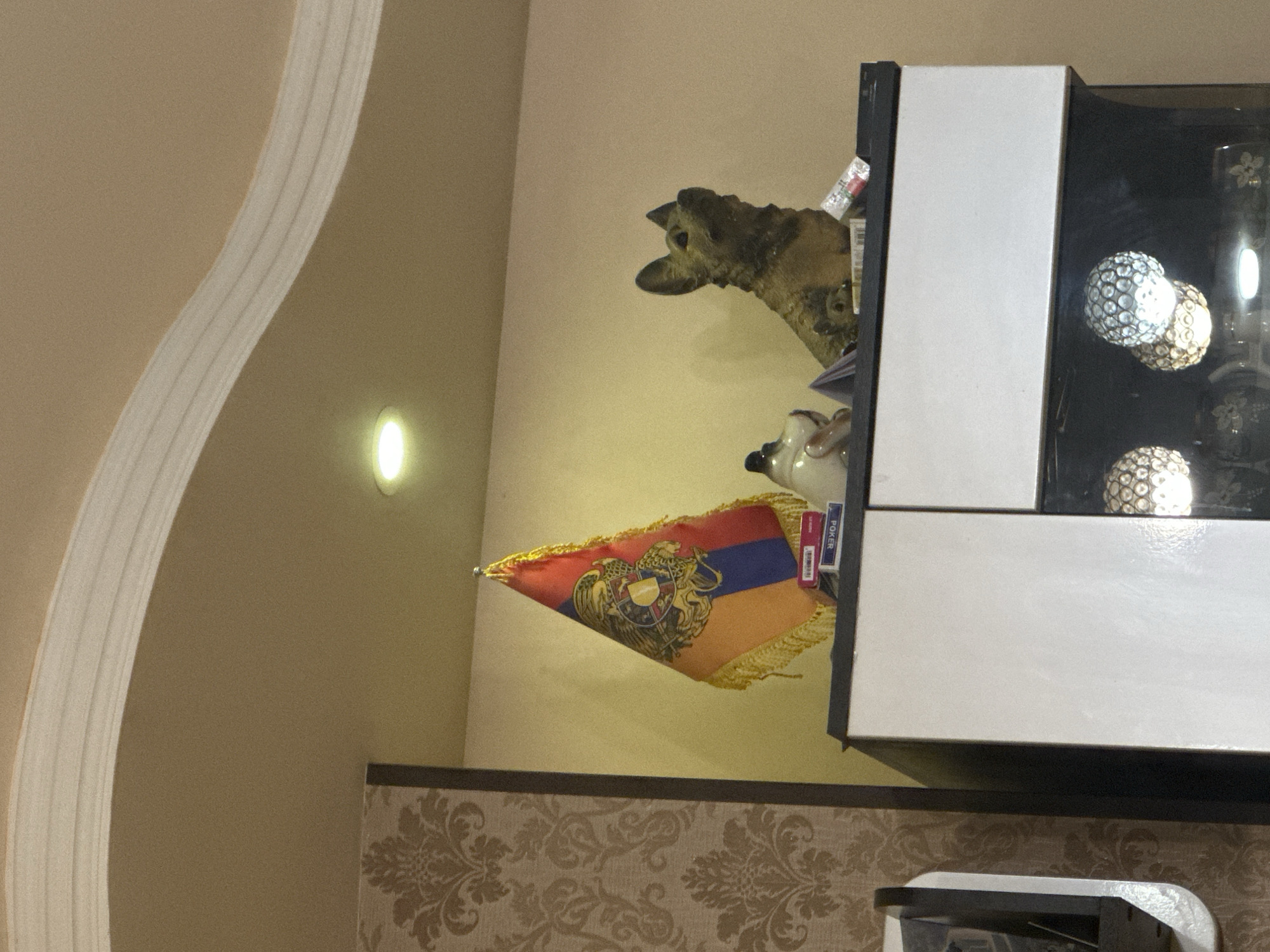 An indoor scene is depicted, focusing on a corner of a room. The walls are a warm, light brown or beige color. On the left, a decorative, white, curved molding ascends the wall. A wall-mounted light fixture casts a soft, yellow glow further down on the left.
In the lower-middle section, a small, triangular flag is prominently displayed. It features the national colors of Armenia (red, blue, and orange) and the Armenian coat of arms in the center, bordered by gold fringe. Above and slightly to the right of the flag, part of a ceramic or carved animal head, possibly a dog or wolf, protrudes from behind a piece of furniture.
To the right, a multi-sectioned piece of furniture, appearing to be a display cabinet or shelving unit, dominates the right side of the frame. It has contrasting white and dark sections. Inside the dark sections, several spherical decorative objects, embellished with small, reflective facets or rhinestones, are visible, illuminated by an internal light source.
On top of this furniture, next to the animal head, several small boxes are stacked. One box clearly displays the word "POKER" in white letters on a blue background, with some additional red text below it that is less legible. The bottom portion of the image reveals a patterned wallpaper in shades of brown and beige, featuring an ornate, damask-like design. There is no discernible activity taking place, and the lighting suggests an indoor setting, possibly during evening hours.
