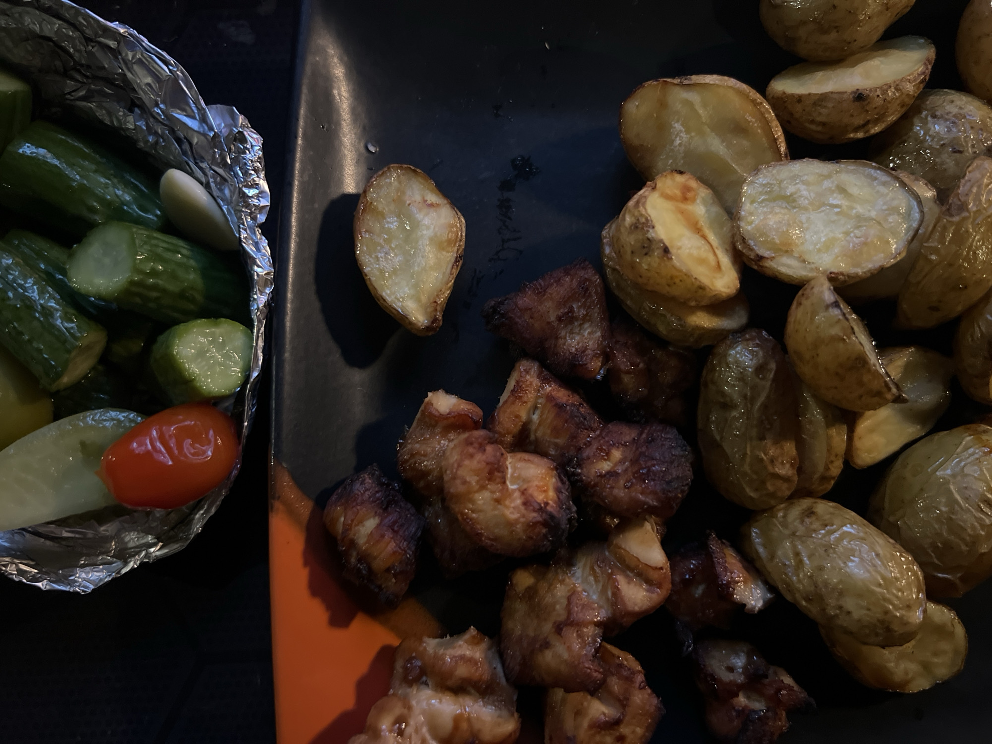 The image shows a close-up of food served on two containers. On the left is a foil container holding sliced cucumbers, a tomato, and a clove of garlic. On the right is a black plate with halved and roasted potatoes and pieces of cooked meat. The location is Sukawati, Indonesia.
