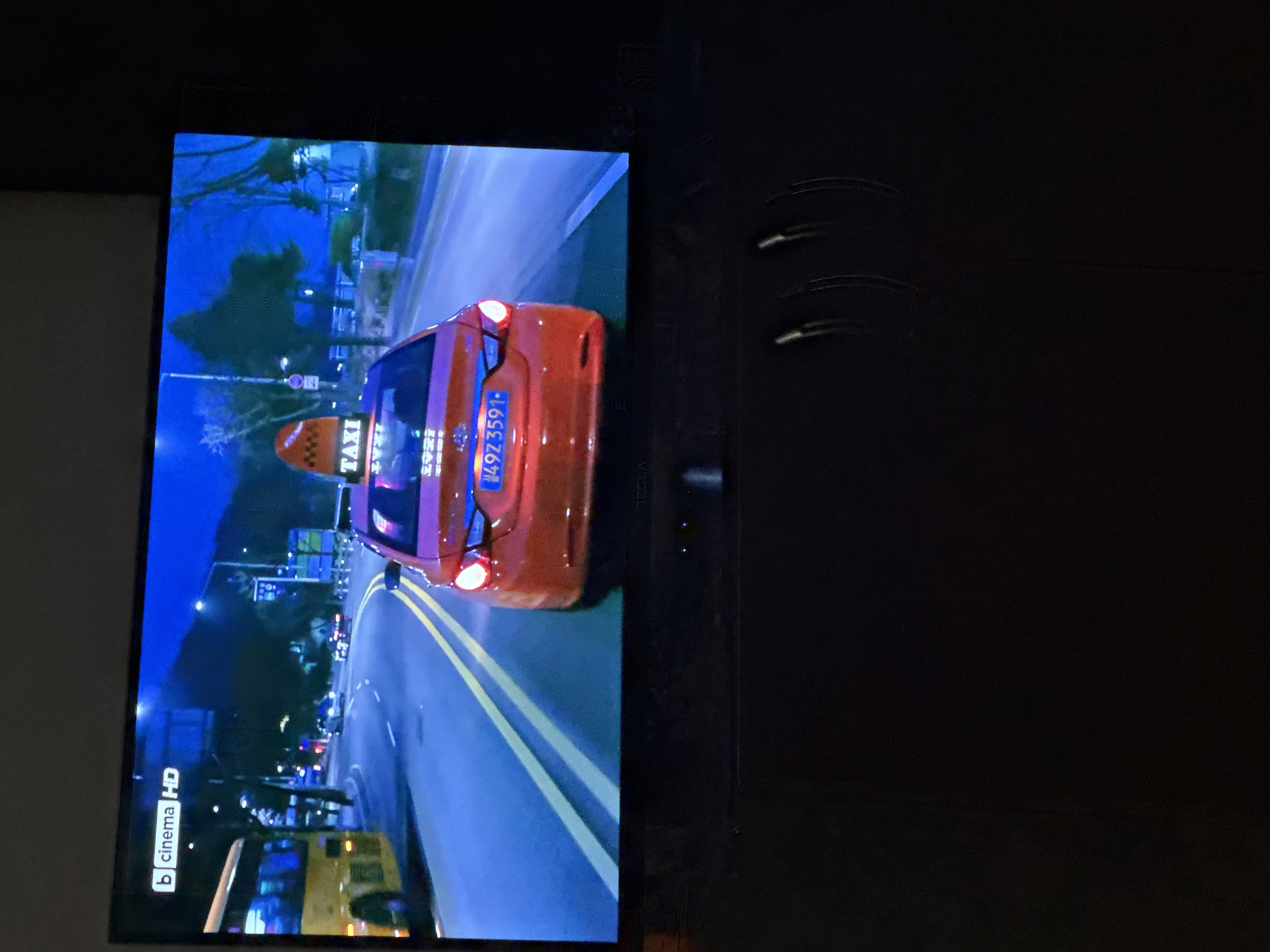 The image depicts a television screen displaying a scene from a film or television show. The scene appears to be set in a city at night, featuring a taxi driving along a road with other vehicles. The taxi's license plate reads "49Z3591". The screen also displays a "b cinema HD" logo. Without additional visual information, it is impossible to determine whether this is a location in Sofia, Bulgaria.