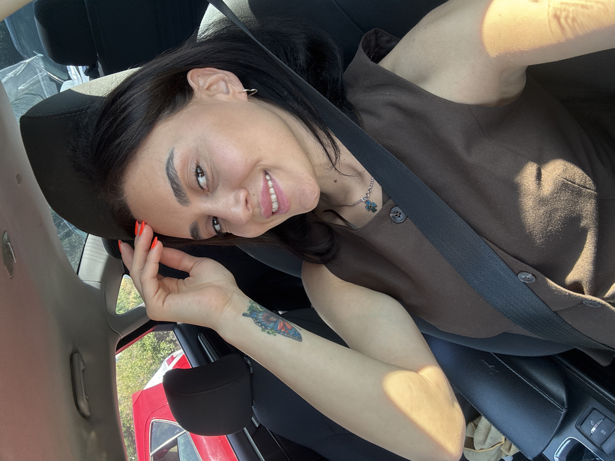 The image shows a woman in the driver's seat of a car. She is smiling at the camera and has her hand on her forehead. She is wearing a brown sleeveless top and a silver necklace with a blue pendant. She has a tattoo of a butterfly on her left forearm. The car interior is visible, including the dashboard, steering wheel, and car seats. Outside the car, another red car and trees are visible. The lighting suggests it is daytime. The location is not specified in the image or provided context.