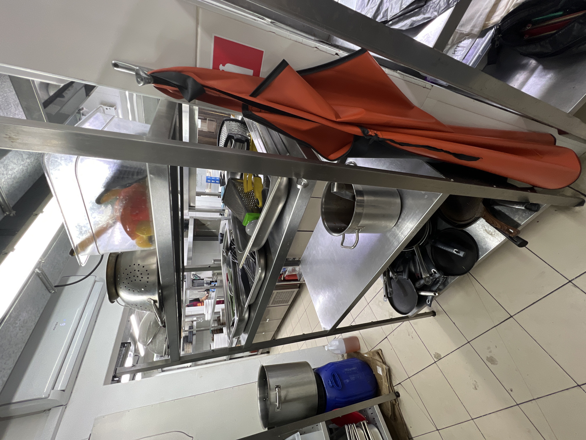 The image depicts a commercial kitchen environment, likely in Plastunivska, Russia, characterized by stainless steel shelving units laden with various culinary equipment. On the top shelf, a metal colander sits to the left of a clear plastic container holding red and yellow objects, possibly fruits or vegetables. Below this, another shelf holds an array of stainless steel trays and utensils, including graters and spatulas. A large stainless steel pot rests on a blue plastic barrel to the left of the shelving unit. On a lower shelf, several black frying pans are stacked, and a stainless steel pot with a scoop is visible. An orange apron with black trim is hanging from a hook on the right side of the shelving unit, next to a red fire extinguisher symbol. The floor is tiled, showing some scuff marks. The overall impression is of a functional, somewhat cluttered, but organized kitchen space.