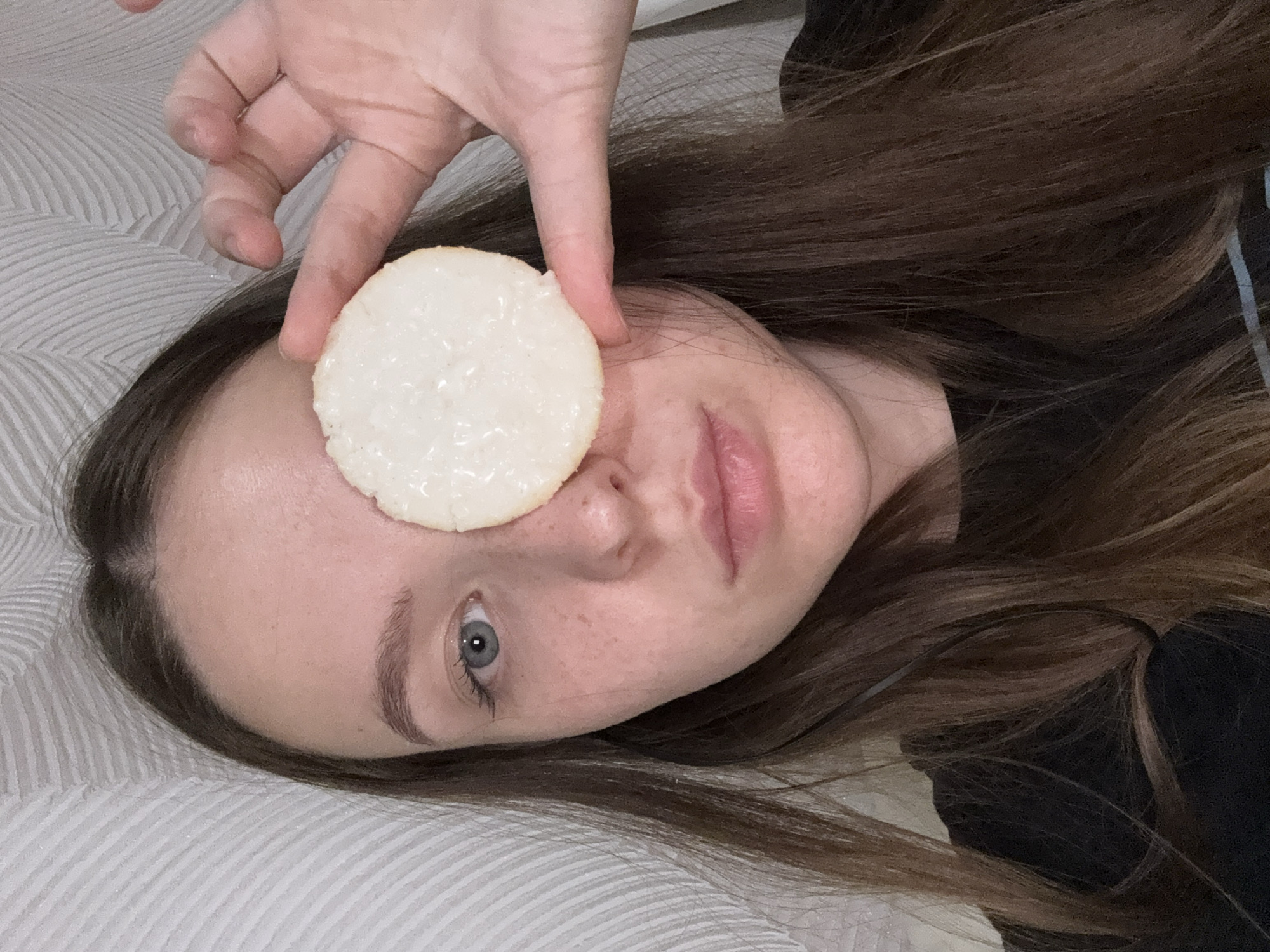 A young woman is depicted in a medium close-up shot, lying down indoors. Her head is tilted, and her left eye, which is blue-grey, looks directly towards the camera. Her right eye is obscured by a flat, circular, light-colored object with a textured surface, which she holds in place with her right hand. This object could be a biscuit, cracker, or a beauty product disc.
The woman has long, dark brown hair that spreads around her head. She has fair skin, with visible freckles on her face, and her lips are slightly parted. A hint of dark clothing is visible near her neck and shoulder.
The background features a light-colored, possibly white or light grey, fabric with subtle wavy or striped patterns, indicative of bedding such as a bedsheet, duvet cover, or pillow. The lighting is soft and even, consistent with an indoor setting, and suggests a casual moment or a self-portrait. No text is visible in the image. The overall impression is one of relaxed playfulness or whimsy.