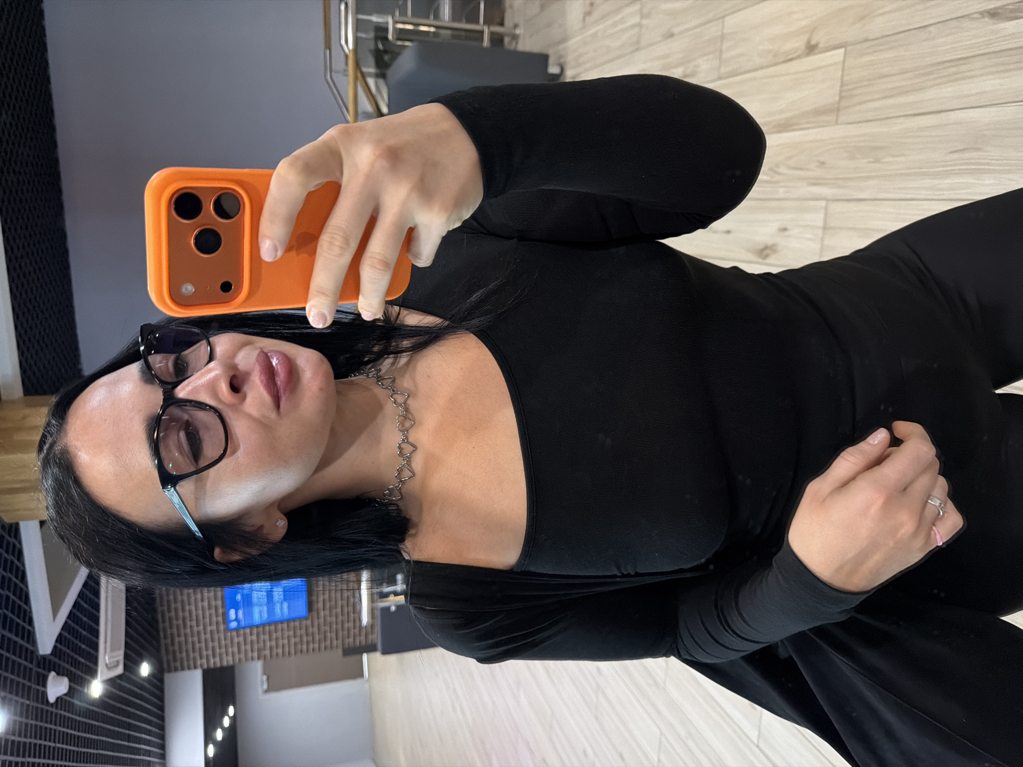 A woman is taking a selfie indoors. She is wearing a black long-sleeved shirt and black glasses. She is holding an orange phone. She is wearing a silver necklace with heart-shaped links. She has a ring on her left ring finger. The background includes a tiled wall, a television screen, and wooden floors. It is not possible to determine if the location is in Saint Petersburg, Russia.