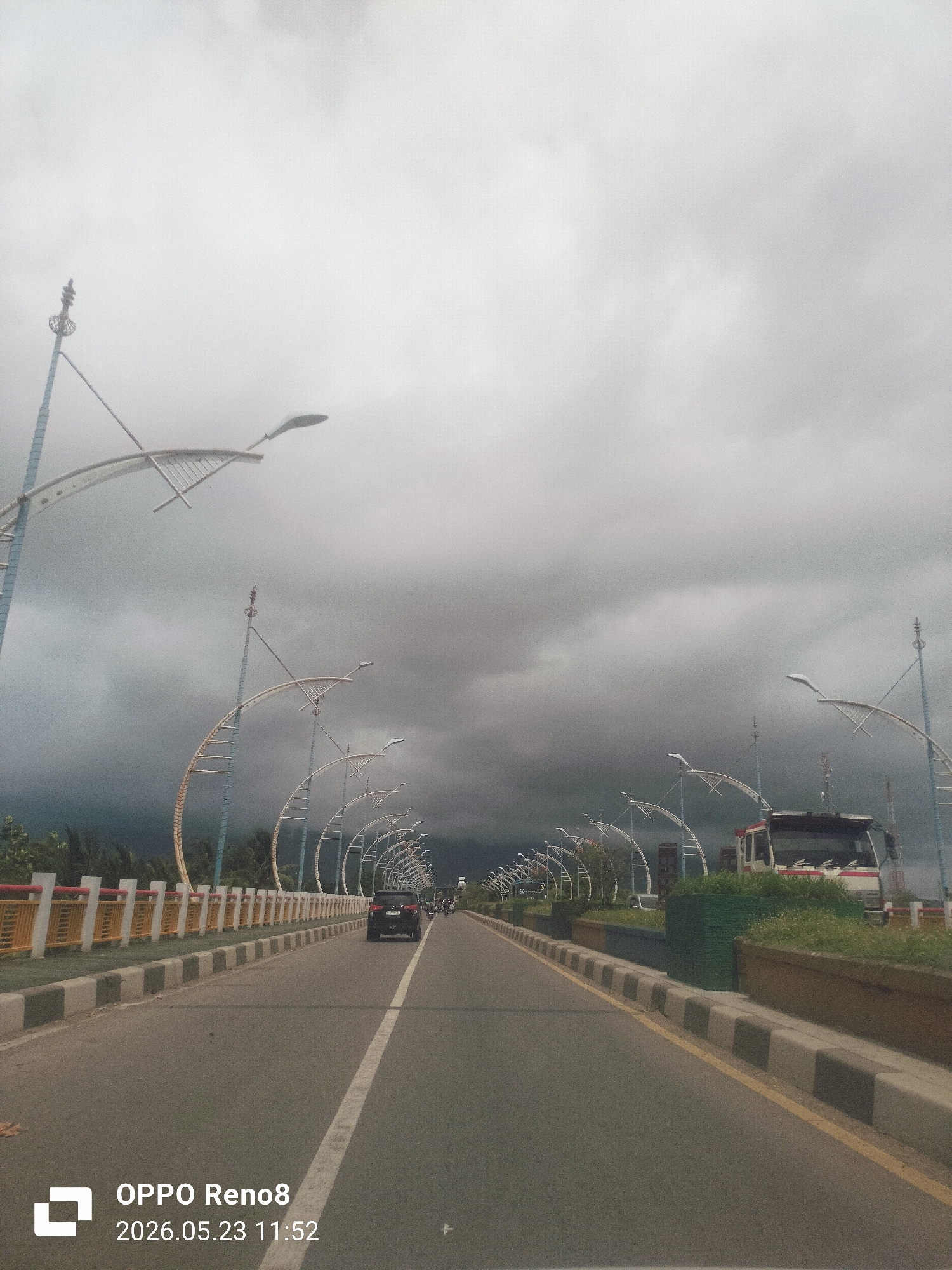 The image shows a road bridge in Lambaro, Indonesia, under a cloudy sky. The bridge has a center white line and bordered sides. On the bridge, there is a car, a truck and other vehicle are visible. Ornamental light fixtures are mounted along the bridge sides. The timestamp "2026.05.23 11:52" is at the bottom left.