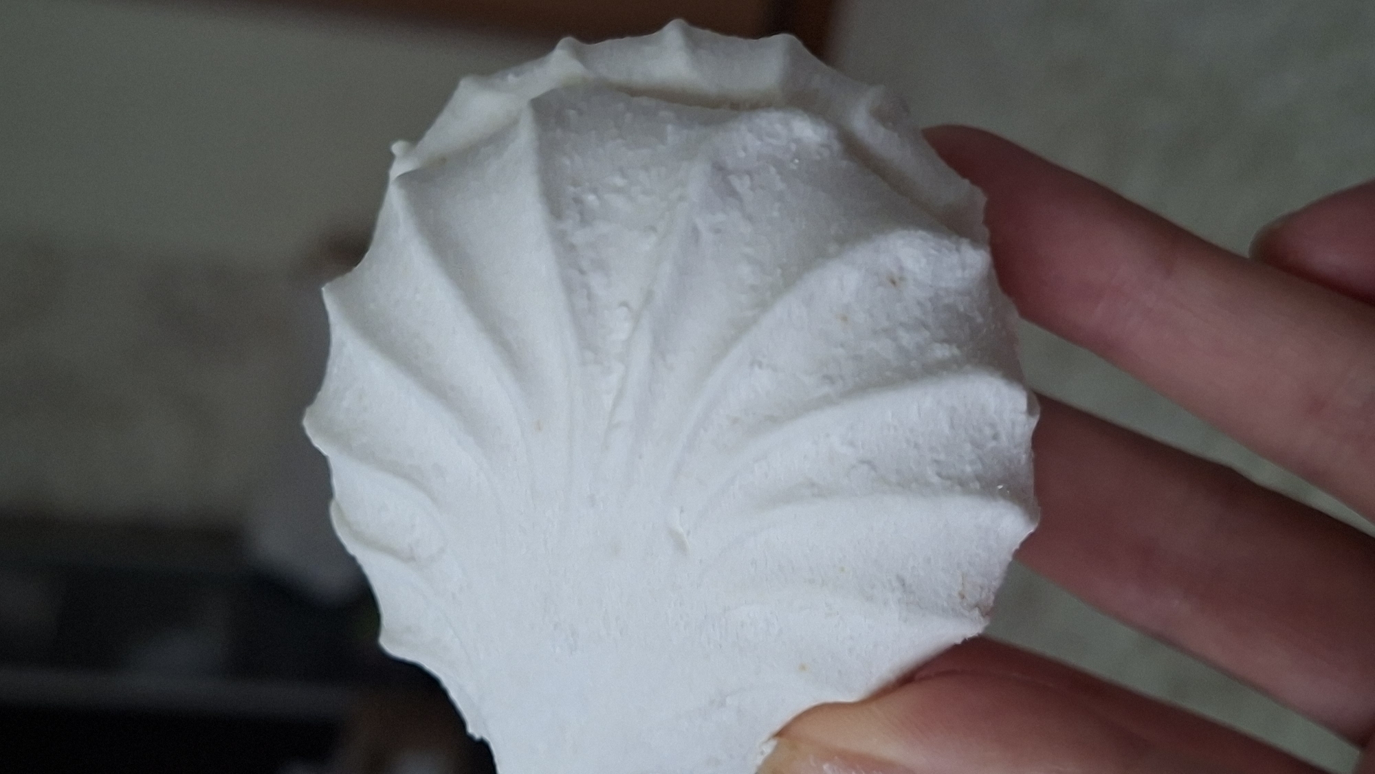 A person's hand is holding a white, ridged pastry, possibly a marshmallow or meringue, against a blurred background. The pastry has a spiral pattern and a textured surface. The hand appears to be that of an adult. The environment is indoors, with the background suggesting a room. The image does not contain any information that could identify its location in the city of Myadzyel, Belarus.