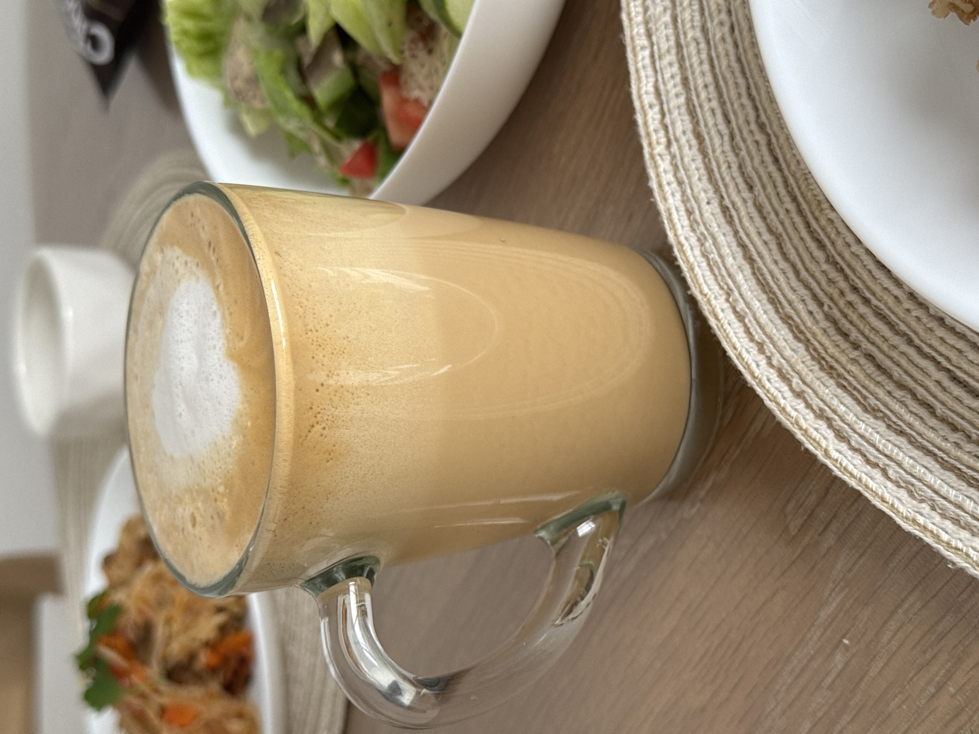 A close-up, eye-level shot captures a clear glass mug filled with a frothy, caramel-colored beverage, likely coffee or a latte, placed on a wooden table. The mug's handle is visible on the left. In the blurred background, to the left, is a white plate with food, possibly noodles and vegetables. To the right, a white bowl contains a salad with green lettuce, cucumber, and tomato. The scene is set in a dining environment, though no specific location in Sakmary, Russia, can be determined from the visual information.