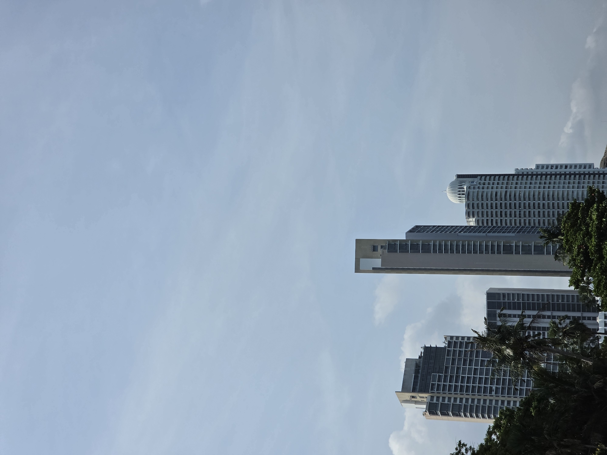 The image depicts a partial view of Pattaya City, Thailand, showcasing several modern high-rise buildings against a bright blue sky. A mix of residential and commercial towers are visible, varying in height and architectural style. The buildings are constructed with glass and concrete, featuring grid-like patterns and horizontal balconies. The foreground contains trees that partially obstruct the view of the lower portions of the structures. The sky shows hints of clouds.