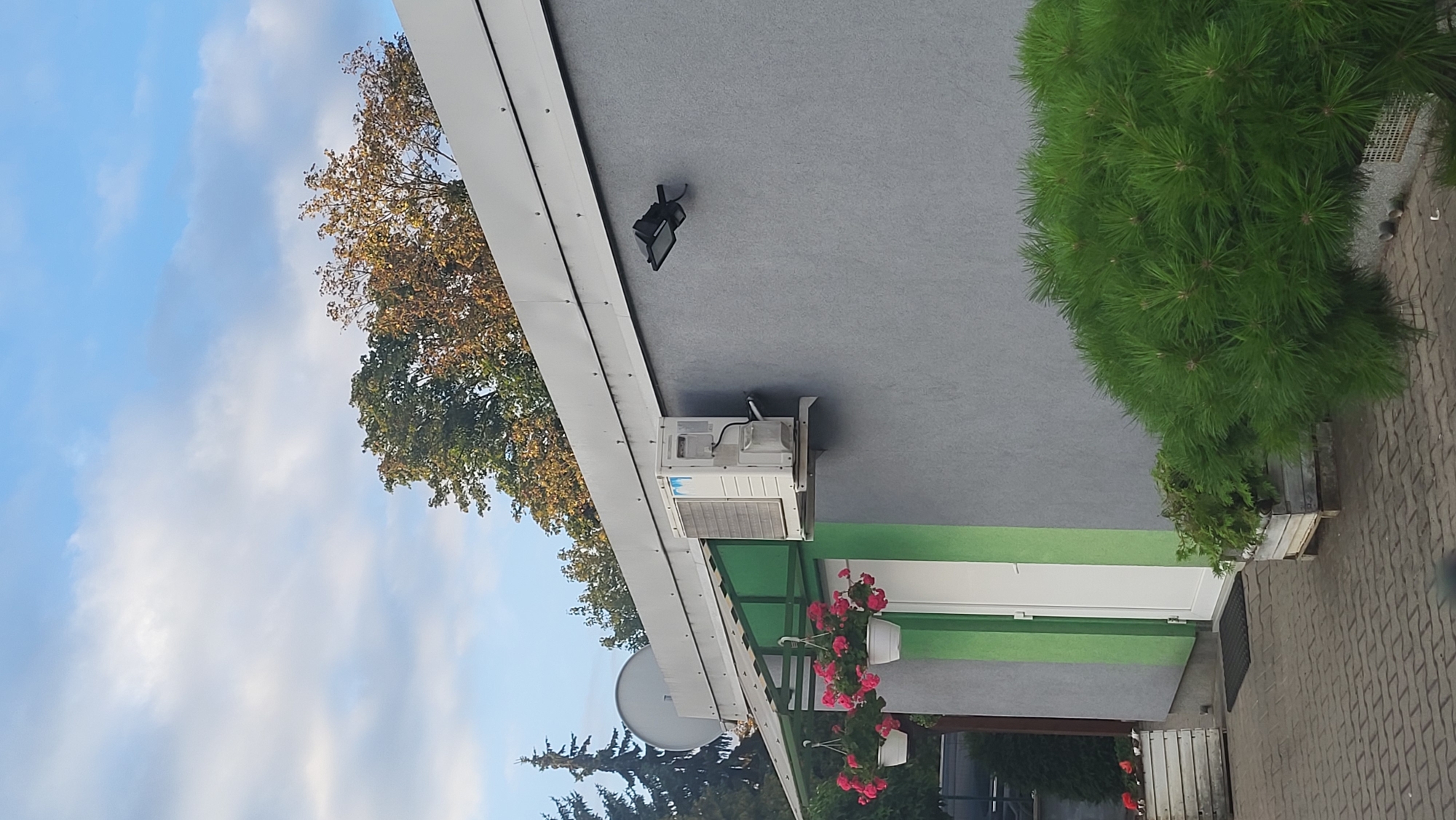 The image captures the exterior of a building with a grey facade, featuring a white door with a green frame. An outdoor air conditioning unit is mounted on the wall, and a black outdoor light fixture is also visible. Several hanging flower pots with pink flowers adorn the overhang above the entrance. A large evergreen bush is situated in the foreground, with a paved area in front of the building. Trees with a mix of green and autumnal foliage are visible in the background, under a partly cloudy sky. The setting appears to be a residential or commercial area in Prešov, Slovakia. No people are present in the image.
