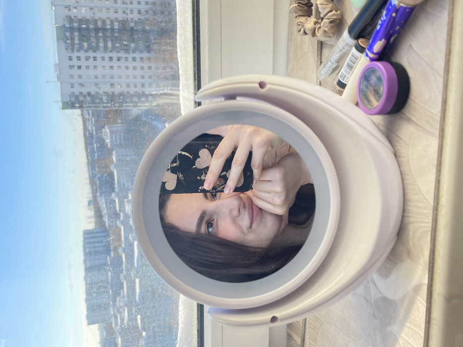 The image captures an indoor scene in Moscow, Russia, looking out onto a snow-covered urban landscape under a clear blue sky. A light pink, circular vanity mirror with an illuminated bezel occupies the foreground, resting on a light-colored windowsill. The mirror reflects the upper left portion of a young woman's face with dark hair, showing a slight smile. Her right hand, holding a smartphone in a dark, heart-patterned case, partially obscures her face while her left hand rests beneath her chin. Adjacent to the mirror, various cosmetic items, including a "BROW GEL SCULPTING" tube, other makeup tools, a purple-lidded container, and several beige scrunchies, are arranged. The background reveals a panoramic view of a residential district with numerous multi-story apartment buildings and a distinct white high-rise with green trim on the right, all covered in snow with visible bare trees, characteristic of a Moscow winter.