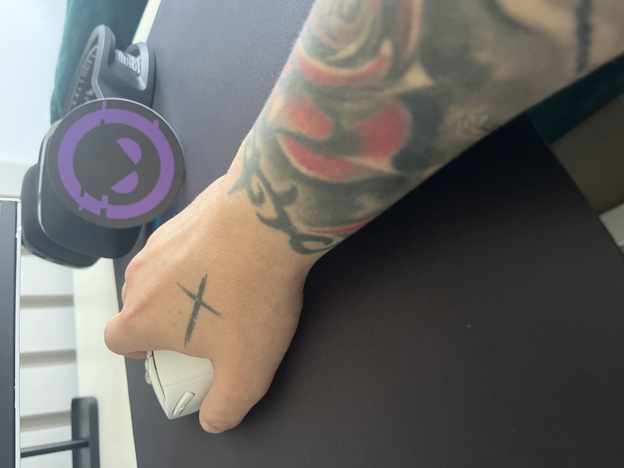 A person's hand with a cross tattoo is holding a white computer mouse on a dark-colored mousepad. The person has an extensive arm tattoo featuring floral designs with red accents. A pair of headphones with a purple circular logo are situated to the left. The background includes a glimpse of a green cushion or piece of furniture and a section of what appears to be a computer tower. The location in Varna, Bulgaria is not discernible from this image.