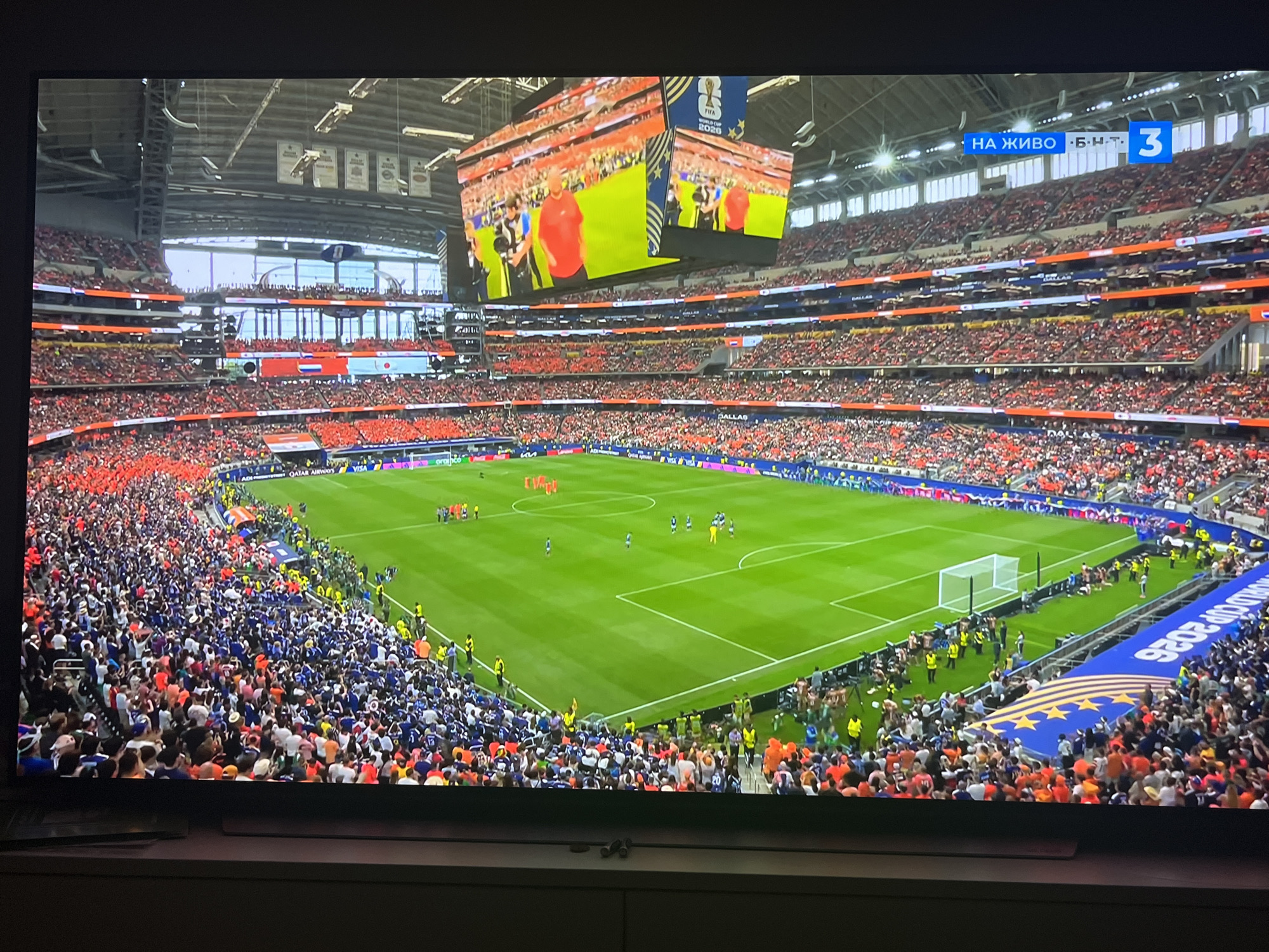 The image captures a wide-angle view of a soccer stadium, likely during a match. The stadium is filled with spectators dressed in predominantly red and blue attire, creating a vibrant atmosphere. The green pitch is clearly visible, with players in orange and blue jerseys engaged in the game. A large scoreboard hangs above the field, displaying a live broadcast of the match and game statistics. In the upper right corner, a banner for "FIFA WORLD CUP 2026" is visible, suggesting the event is a qualifying match or promotional event related to the upcoming World Cup. The overall scene is dynamic and energetic, indicative of a live sporting event. There is no indication of Varna, Bulgaria in the image.