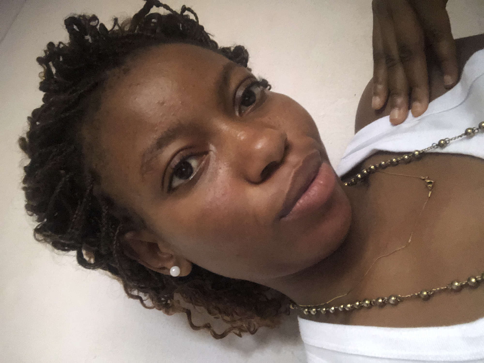A close-up selfie of a woman with dark brown skin, dark brown locs, wearing a white top and a gold necklace with beads. She is wearing a pearl earring. The background is a plain white wall.
The city of Kashere, Nigeria is not discernible in the image.