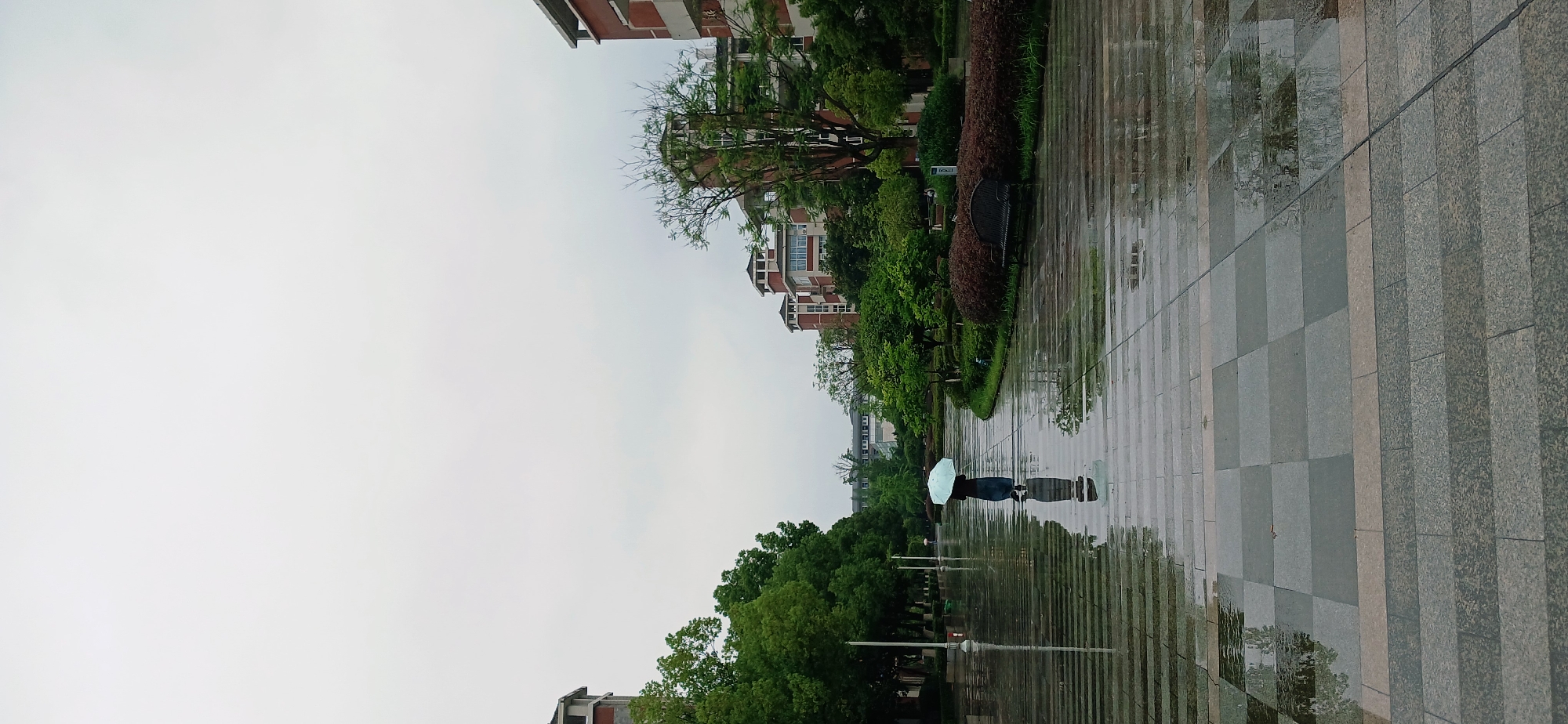 A lone individual walks away from the camera, holding a light blue umbrella. They are on a wet, paved pathway that reflects the overcast sky and surrounding buildings. The pathway is laid with rectangular tiles of varying shades of gray and beige. To the left of the pathway are trees and a building. To the right, there is a manicured garden area with shrubs and a bench, and beyond that, more buildings with red brick exteriors and white trim. The sky is uniformly gray, suggesting rainy weather. The scene appears to be a campus or park-like setting within the Chongqing High-tech Zone, China.