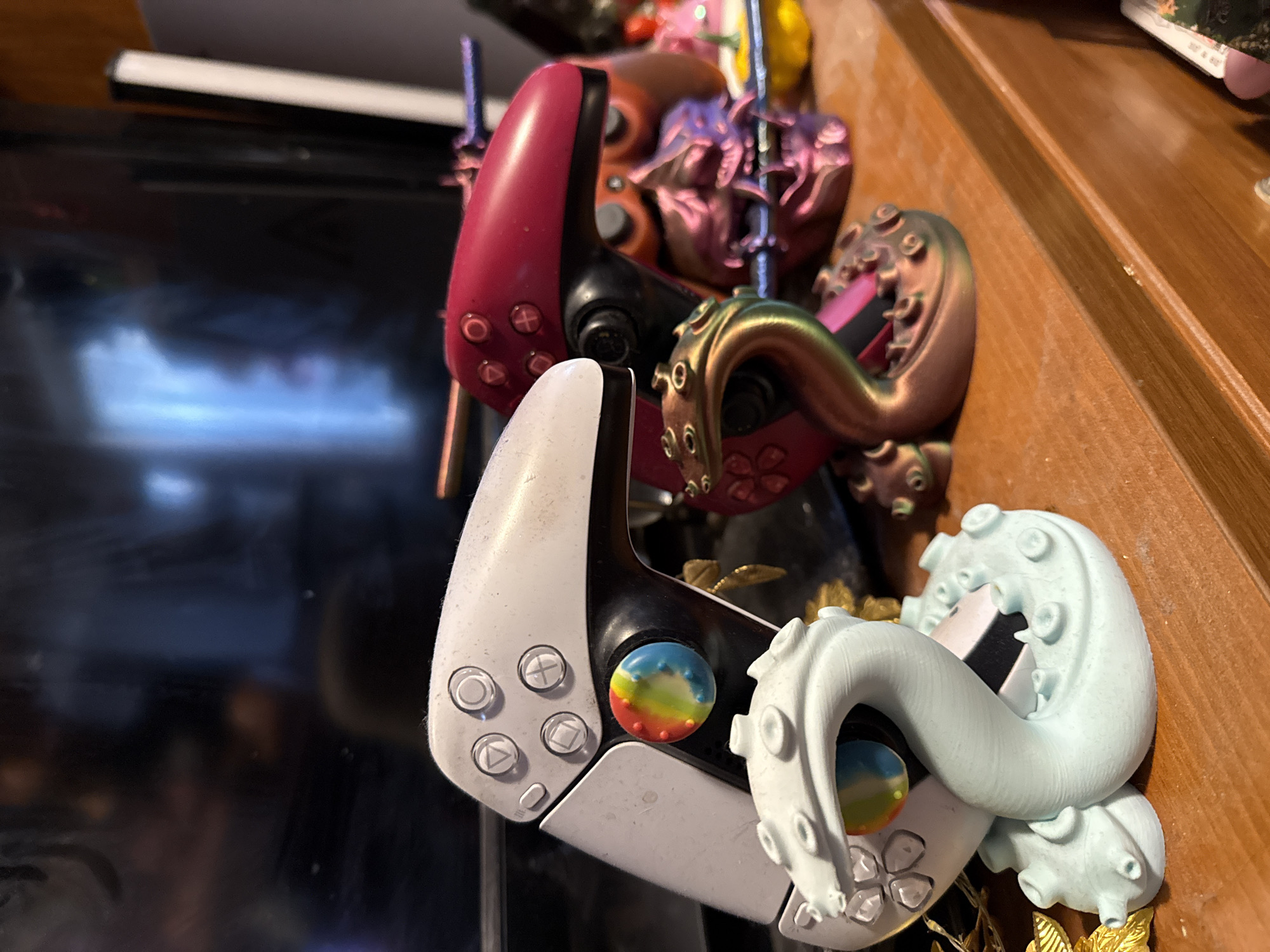 The image shows several PlayStation 5 DualSense controllers, customized with decorative elements such as tentacles. The controllers are arranged on a surface, partially obscuring each other. The controller closest to the viewer is white, adorned with a light blue tentacle. Another controller behind it is predominantly red. The surface they are on appears to be a wooden shelf, and a television screen is visible in the background. The location is not possible to determine from the image.