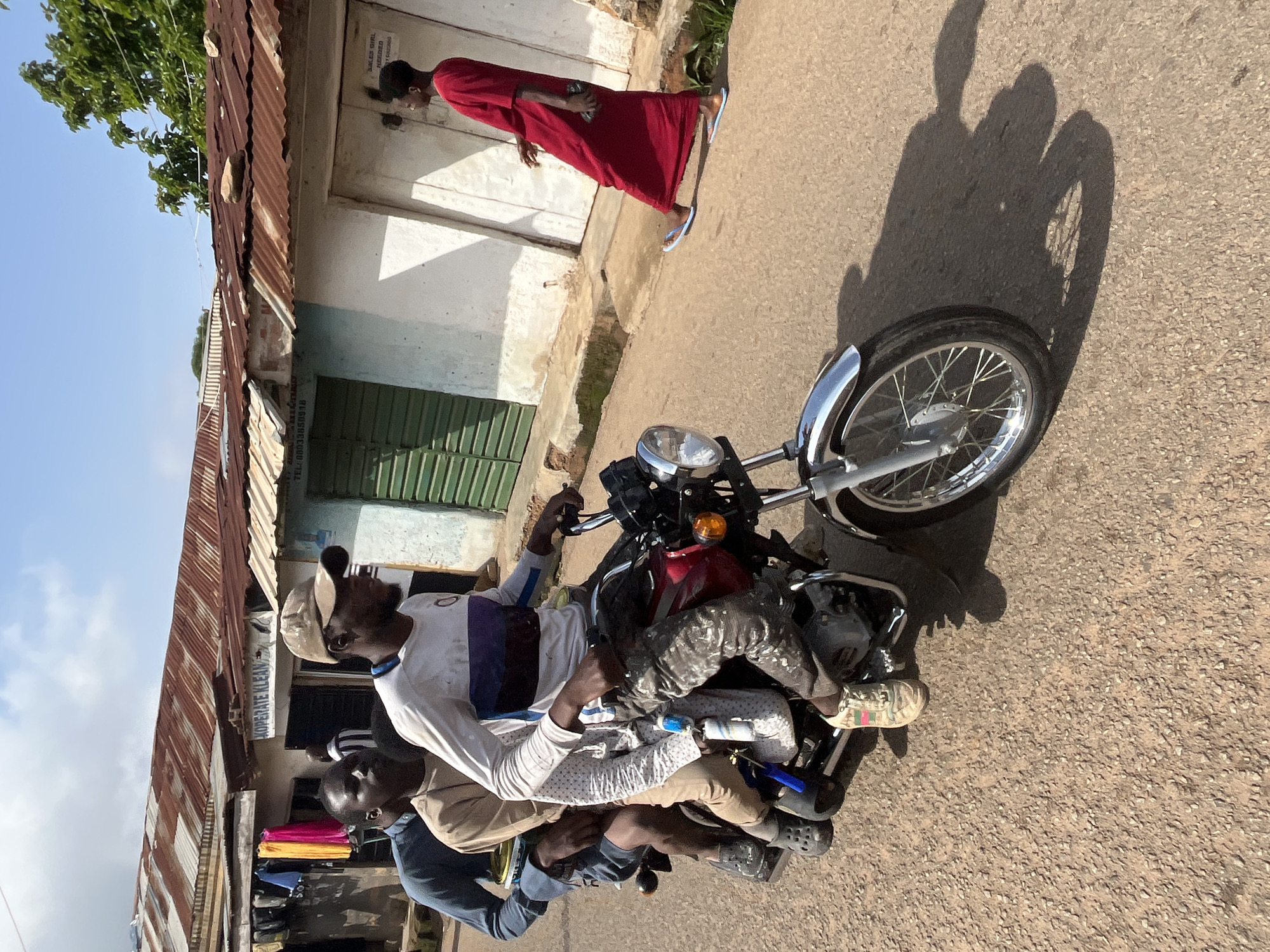 A photograph taken outdoors shows three men on a motorcycle in a street setting in Jos, Nigeria. The man in the front is driving the motorcycle and is wearing a white long-sleeved shirt with horizontal stripes of purple and dark blue, grey camouflage patterned pants, and a tan baseball cap. The second man is in the middle, wearing a tan shirt and black clogs. The third man is in the back, wearing a blue long-sleeved shirt and dark shorts. The motorcycle is red and silver with a visible engine and silver handlebars. In the background, there are buildings with corrugated metal roofs, some of which appear to be shops. One building has a sign that reads "XOPERATE KLEAN." The street is paved with a gravelly surface. A person wearing a long red dress and light blue flip-flops is walking away from the motorcycle on the right side of the street. The sky is visible above with some clouds.