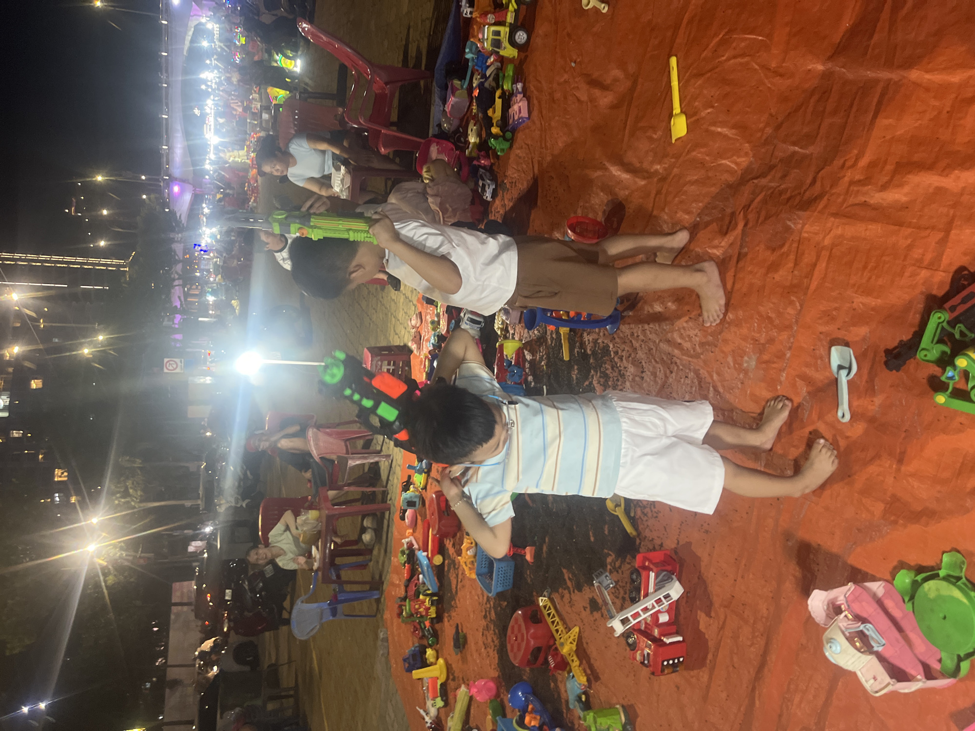 An outdoor scene at night in the city of Đà Nẵng, Vietnam, features a large gathering of people and children amidst a sea of toys spread across orange tarps. The environment is illuminated by numerous bright streetlights and possibly decorative lighting, creating a festive atmosphere. In the foreground, two young boys are actively engaged with toys. One boy, wearing a striped t-shirt and white shorts, is holding a green toy gun to his ear. The other boy, in a white t-shirt and brown shorts, stands behind him, also holding a green toy gun. The ground is covered with a variety of colorful plastic toys, including toy cars, trucks, building blocks, and other playthings. In the background, other individuals are visible, some seated and others standing, suggesting a public event or market. The overall scene depicts a lively and communal gathering, likely a night market or a special event catering to families.