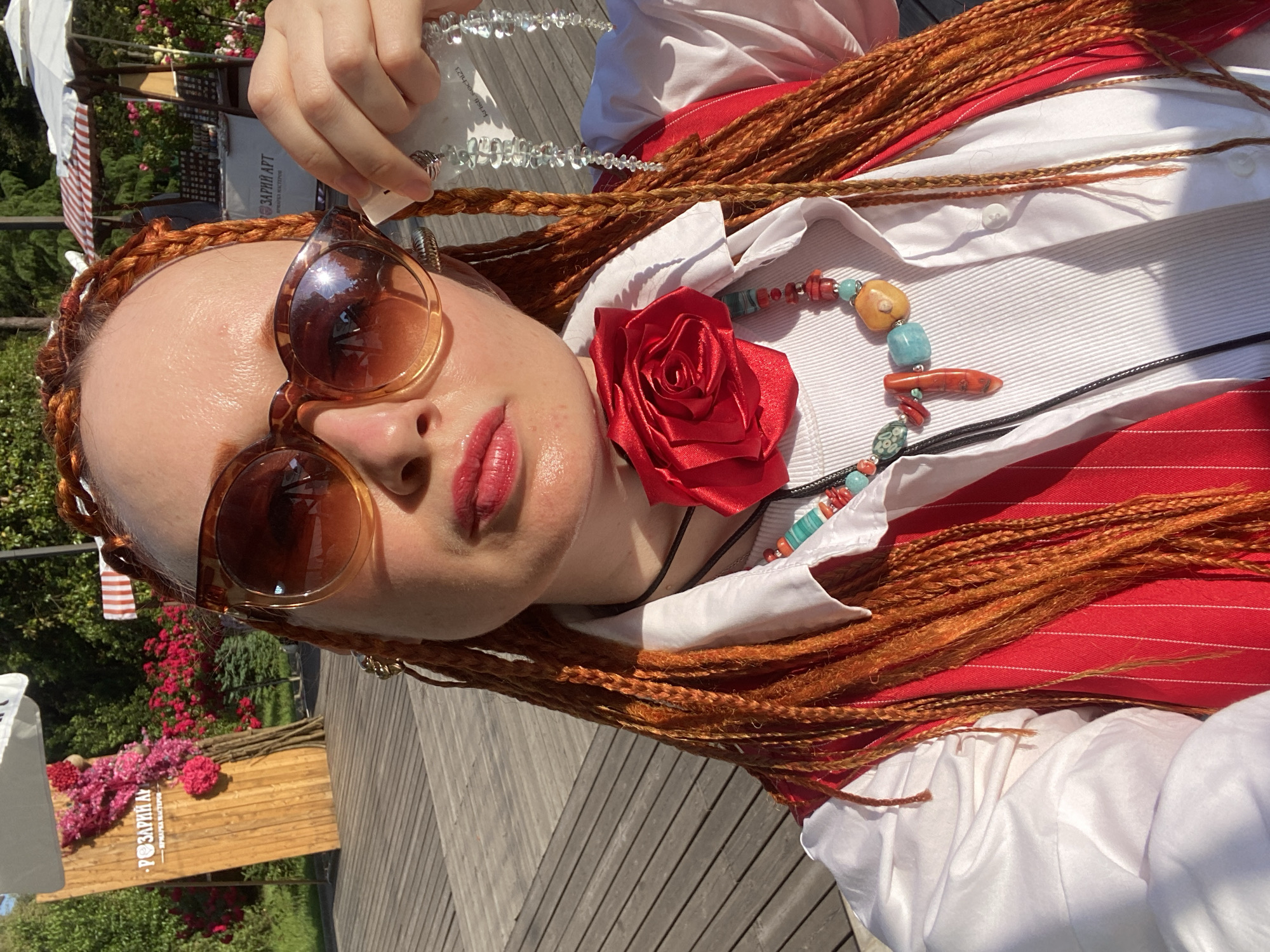 A woman with auburn braided hair and bright red lipstick is taking a selfie in an outdoor setting. She is wearing oversized amber-tinted sunglasses, a white collared shirt, and a red pinstriped vest. Around her neck, she has a statement necklace featuring a large red fabric rose, colorful beads, and a coral-like pendant. She is holding up a long, clear beaded necklace. In the background, there are wooden structures, lush greenery, and red and pink flowers, suggesting a park or garden setting. A sign with Cyrillic text is visible on a wooden board. This scene is likely in Sochi, Russia, given the presence of the Cyrillic sign and the summery outdoor environment.