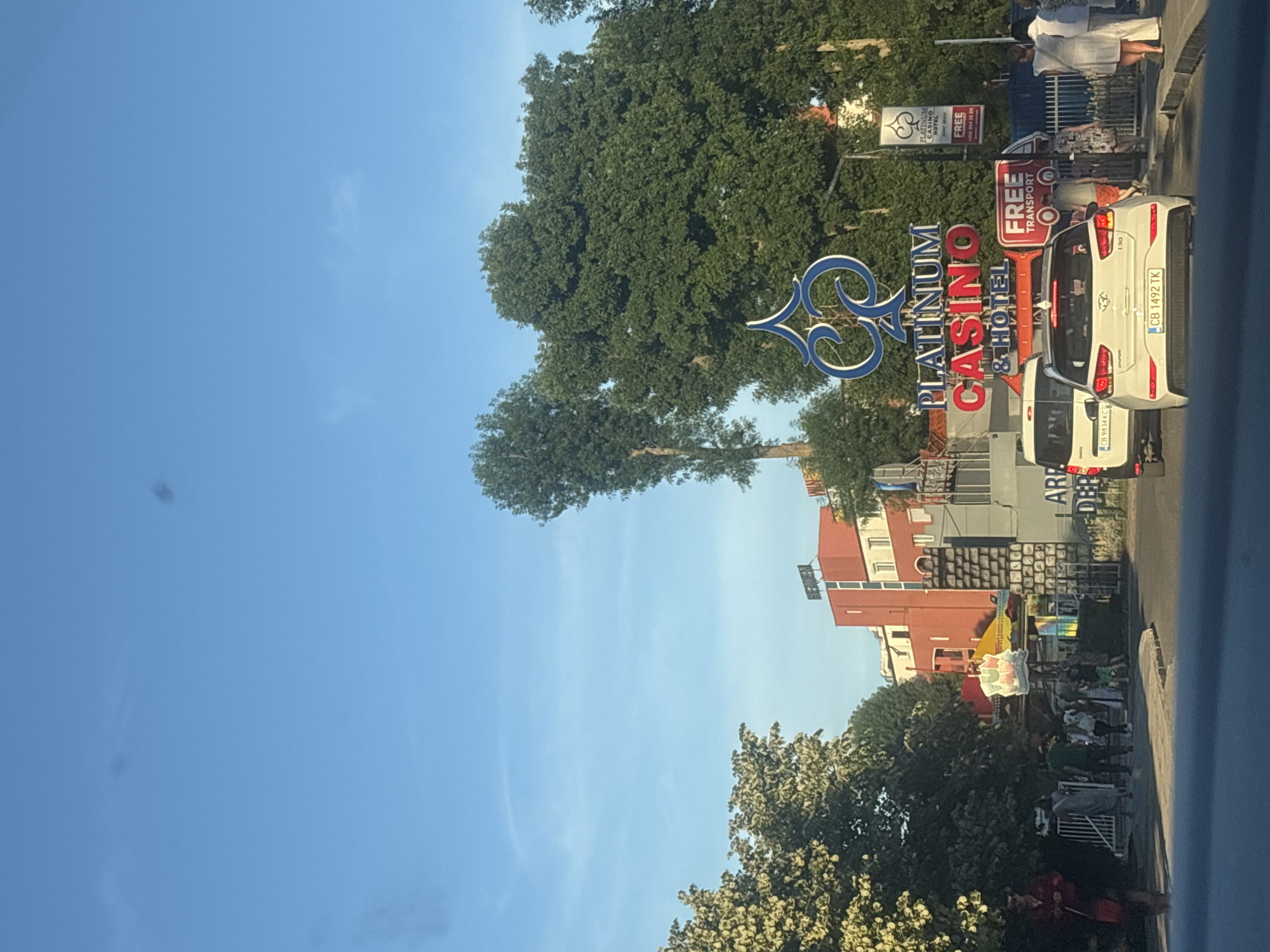 In Nesebar, Bulgaria, a street scene unfolds under a clear blue sky with scattered clouds. Prominently displayed is a large sign for "PLATINUM CASINO & HOTEL" with a distinctive blue spade logo above it. To the right of this, another sign advertises "PLATINUM CASINO MOTEL FREE TRANSPORT." In the foreground, a white Hyundai i30 with a Bulgarian license plate "CB 1492 TK" is stopped on the road, as is a white van with a Bulgarian license plate "CB 9814 KC." Several pedestrians are visible on the sidewalk, some walking and others standing. Behind the casino sign, a red building with white windows stands out, and further back, more buildings and trees are visible. The overall atmosphere suggests a sunny day in a bustling area.