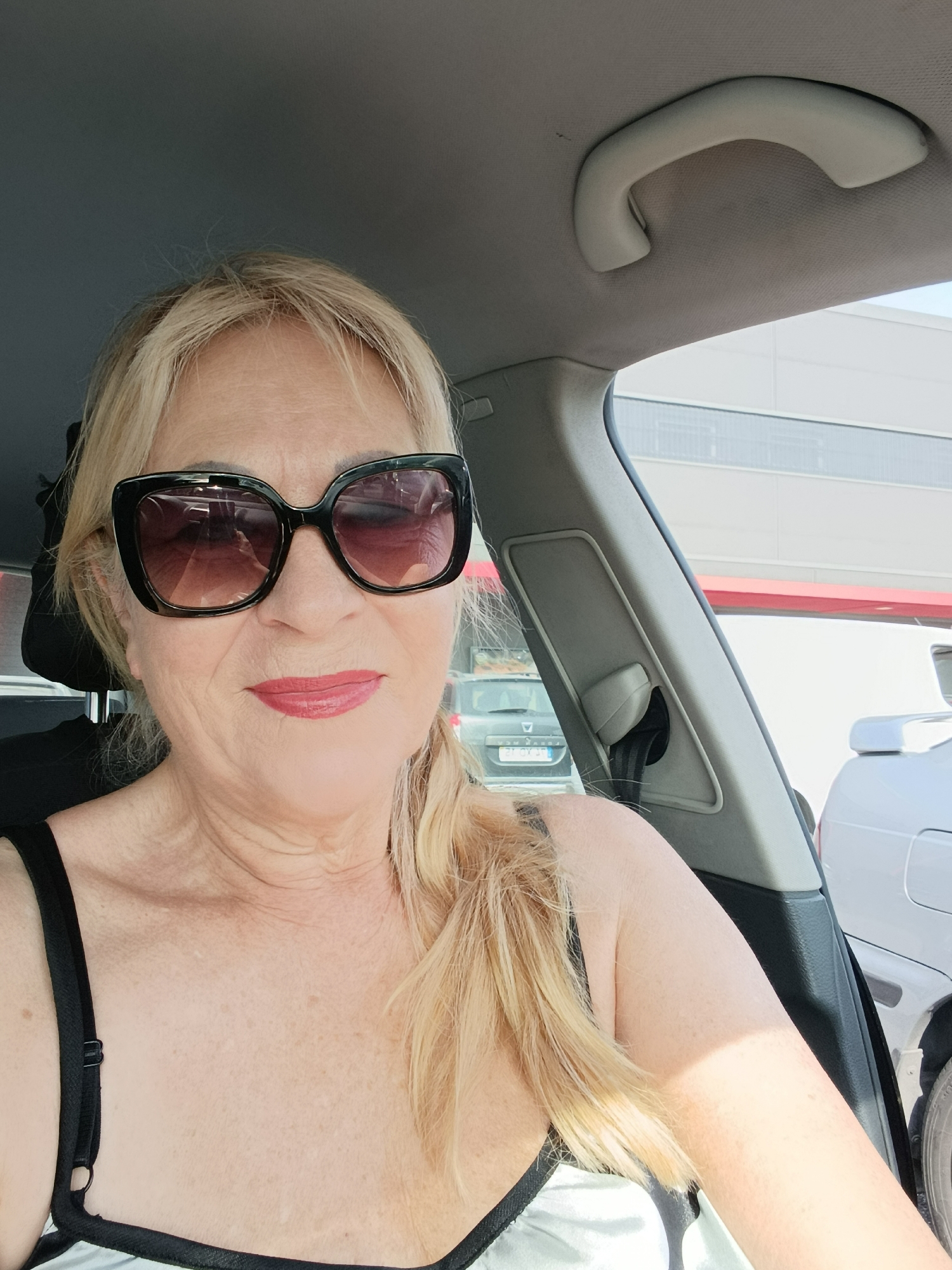 The image shows a middle-aged Caucasian woman with blonde hair, wearing large, black sunglasses and red lipstick, taking a selfie inside a car. The woman is seated in the driver's seat, and her upper body is visible. She is wearing a black bra. The interior of the car is visible, including the grey headliner, a grab handle, and the A-pillar. Outside the car, other vehicles and a building with a red stripe are visible. The image appears to have been taken in Setúbal, Portugal.