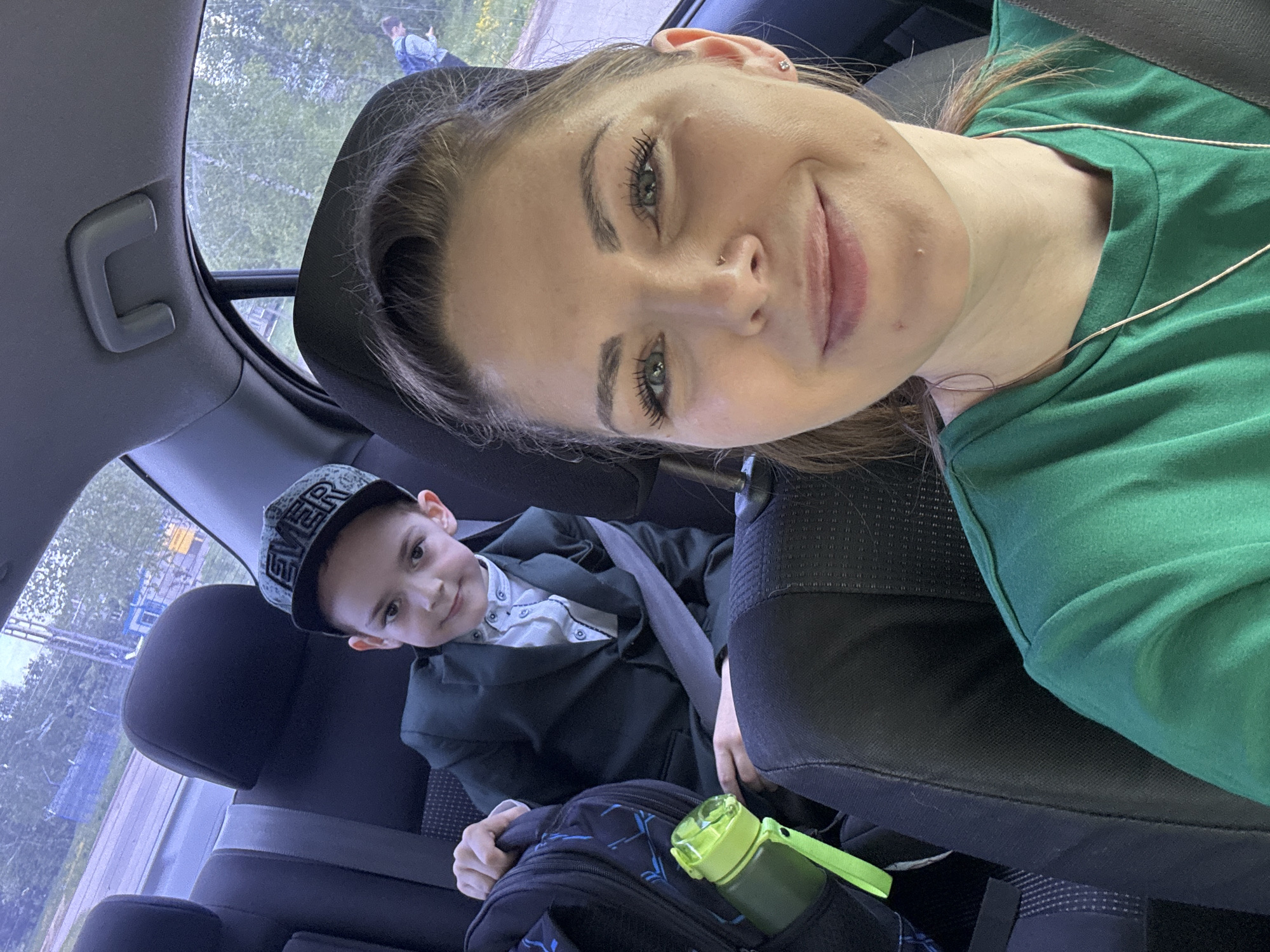 The image is a selfie taken inside a car. An adult female with fair skin, light brown hair, and green eyes, wearing a green shirt and a silver necklace is visible in the front seat. She has a nose piercing and is smiling slightly at the camera.
Behind her, in the back seat, is a young boy wearing a dark blazer, a white button-up shirt, and a baseball cap with the word "EVER" on it. He is also wearing a seatbelt and is looking toward the camera with a slight smile. A backpack and a green water bottle are on the seat in front of him.
Outside the car windows, there is greenery and a glimpse of other buildings suggesting an urban or suburban environment. A person is visible in the distance outside the car.
The image location cannot be determined as Shevlyakovo, Russia.
