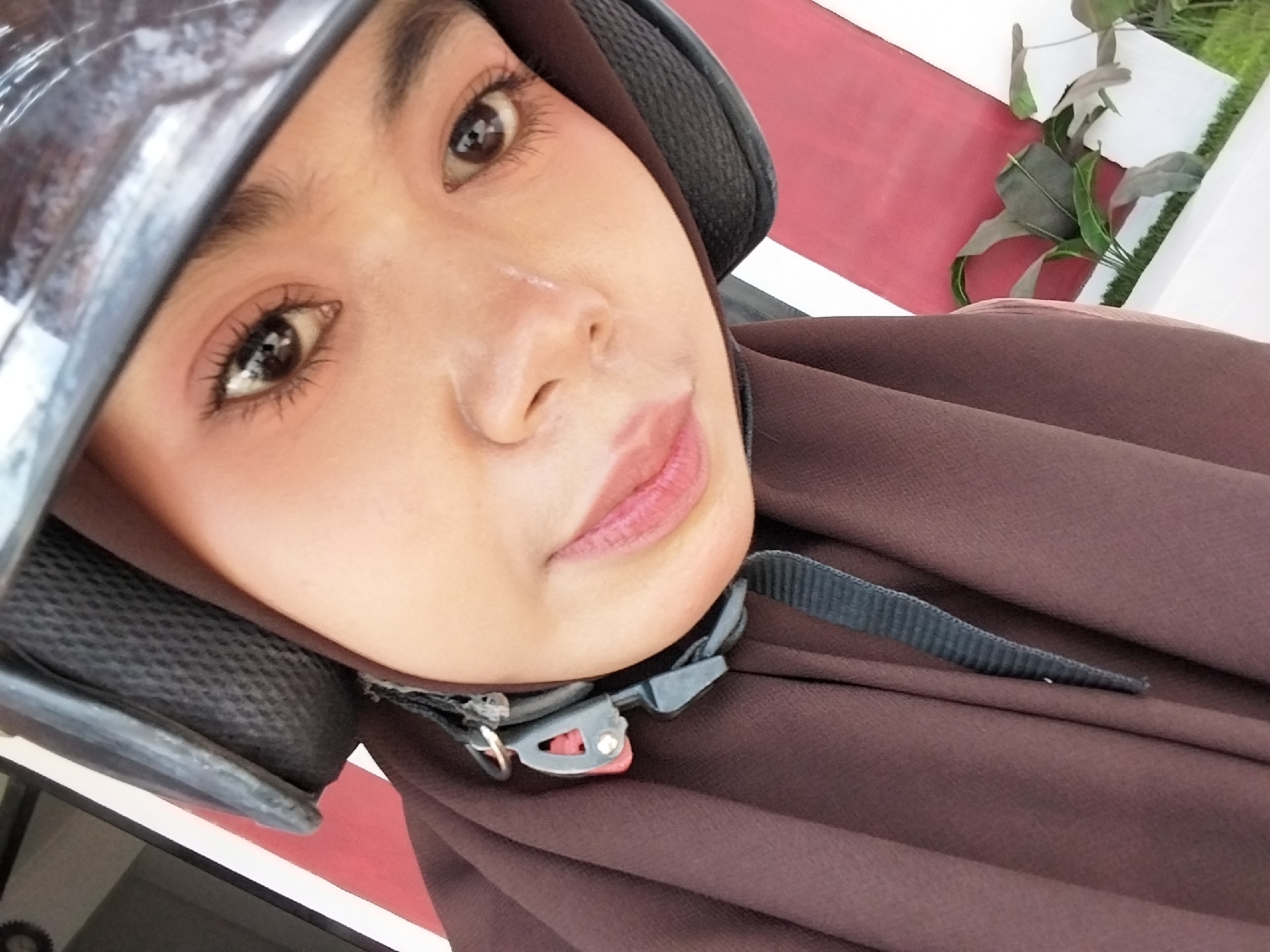 A close-up selfie features a young woman wearing a brown hijab and a black motorcycle helmet. Her face, partially obscured by the helmet and head covering, is angled towards the camera. She is looking directly at the lens with her eyes, which are adorned with noticeable mascara and eyeliner. Her lips are painted a soft pink. The background shows a striped pattern of red and white, with some greenery and what appears to be artificial grass visible in the lower right corner. The image was captured in Tibojong, Indonesia, suggesting an outdoor urban or semi-urban setting.