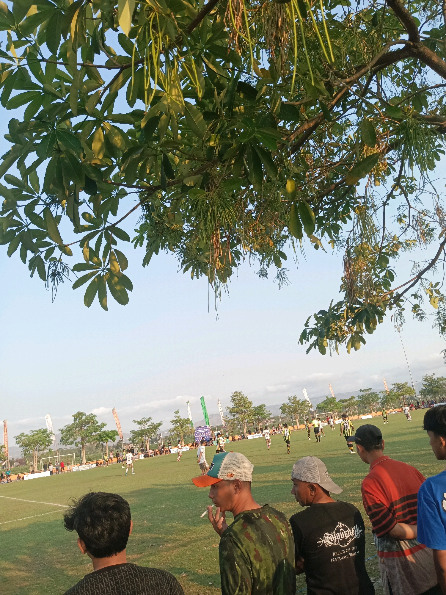 The image depicts an outdoor soccer match on a grassy field, likely in the city of Tawing, Indonesia, given the context. The foreground is dominated by the dark canopy of a tree with long, dangling pods and green leaves, partially obscuring the sky. In the middle ground, several people are visible from behind, spectating the game. One individual in the foreground right is wearing a red and black striped shirt and a black baseball cap. To his left, another person wears a grey baseball cap and a black t-shirt with white lettering. Another spectator in a camouflage shirt is also visible.
The soccer field itself is well-maintained, with white lines marking the boundaries. Several players in yellow and black striped uniforms and white uniforms are actively participating in the game. Goalposts are visible at either end of the field. In the background, rows of trees and distant hills form the landscape. Several flags and banners are erected around the field, suggesting an organized event. The sky is a clear, pale blue, indicating daytime. The overall scene suggests a casual sporting event, possibly a local league match or friendly game.