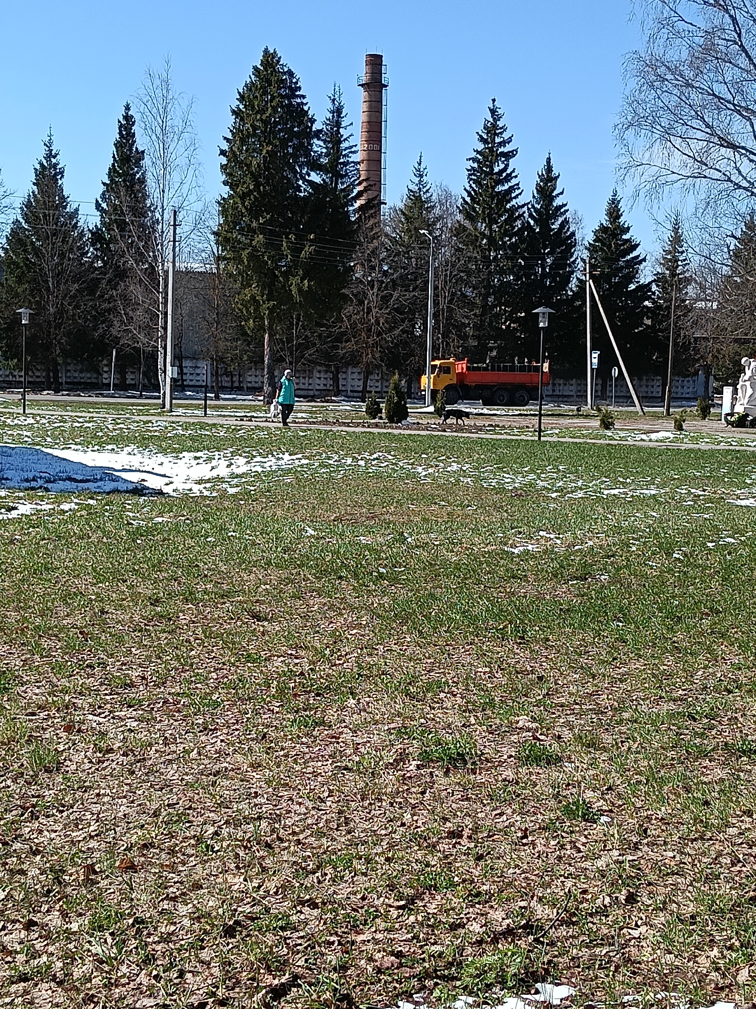 The image displays an outdoor scene in Красный Маяк, Russia, likely during early spring. In the foreground, a large expanse of green grass is visible, partially covered by scattered brown leaves and melting patches of snow.
In the mid-ground, a paved path extends horizontally across the frame. A person, wearing a light blue or green jacket, walks along this path, accompanied by a light-colored dog. Further to the right on the path, another dark-colored dog is also visible. Parallel to the path runs a road where a prominent yellow and orange truck, possibly a utility vehicle or fire truck, is either parked or moving.
The background features a line of tall trees, including numerous dark green evergreens and some deciduous trees with bare branches, consistent with the early spring season. A striking industrial element is a tall, reddish-brown brick chimney or smokestack, on which the numbers "2000" can be faintly observed. Utility poles and power lines are visible against a clear, bright blue sky. Streetlights are positioned along the path and road. A concrete or stone barrier separates the green area from the roadway and structures beyond. A small white statue or monument is discernible in the far background on the right.
The scene is bathed in bright daylight, with a clear sky, suggesting sunny weather. The activity depicted is a tranquil, everyday occurrence of a person walking a dog in a public green space, set against a backdrop that includes both nature and industrial architecture.