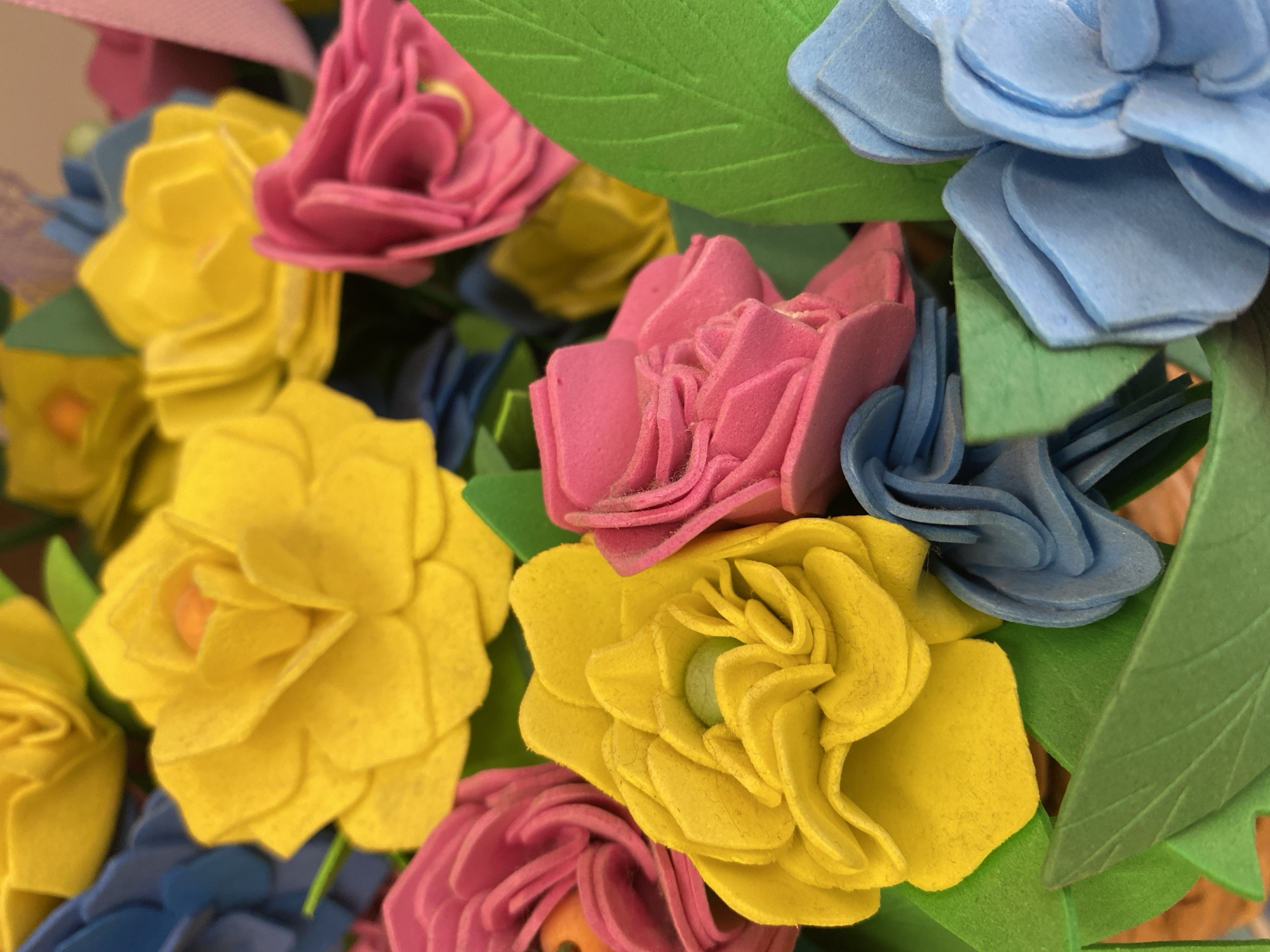 The image shows a close-up of a bouquet of artificial flowers. The flowers are made of felt in various colors: yellow, pink, and blue. There are also green felt leaves scattered throughout the bouquet. The focus is on the textures and colors of the felt materials. The image does not appear to be taken in a specific location in Chaikovsky, Russia, as it is a studio shot or a close-up of an isolated object.
