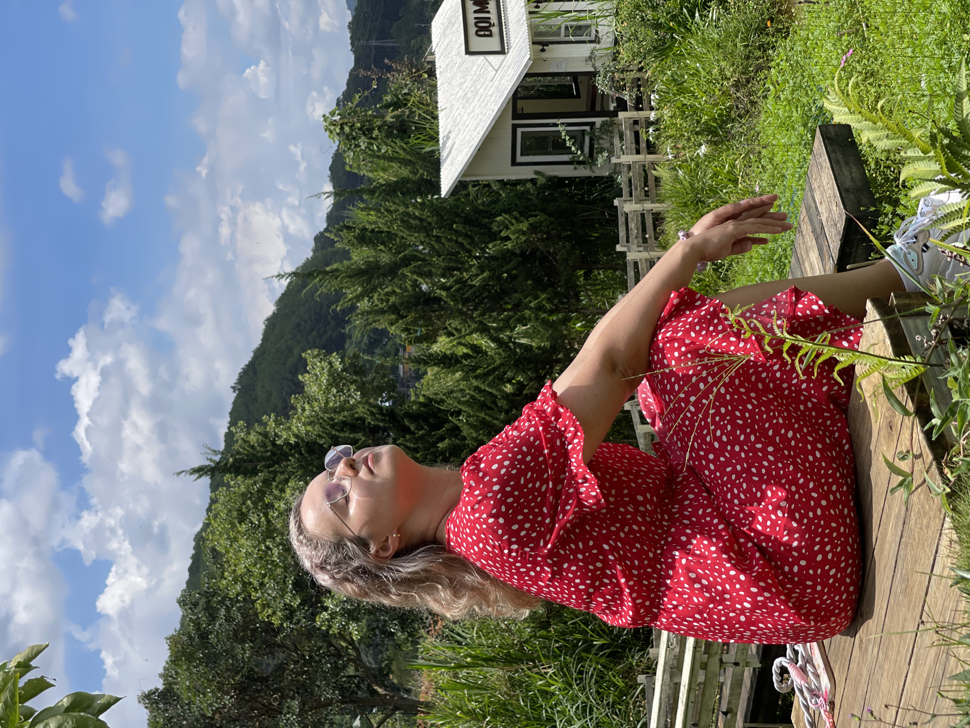 A woman in a red and white polka-dot dress sits on a wooden surface outside, with her head tilted back and eyes closed as she faces the sky. She wears sunglasses and has blonde curly hair. The surrounding environment includes greenery, a small white building with black trim and the text "DOI MƠ" above the door, a mountain range in the background, and a blue sky with scattered clouds. The image is taken at an angle, with the camera positioned slightly to the side and below the woman. The location is Da Lat, Vietnam.
