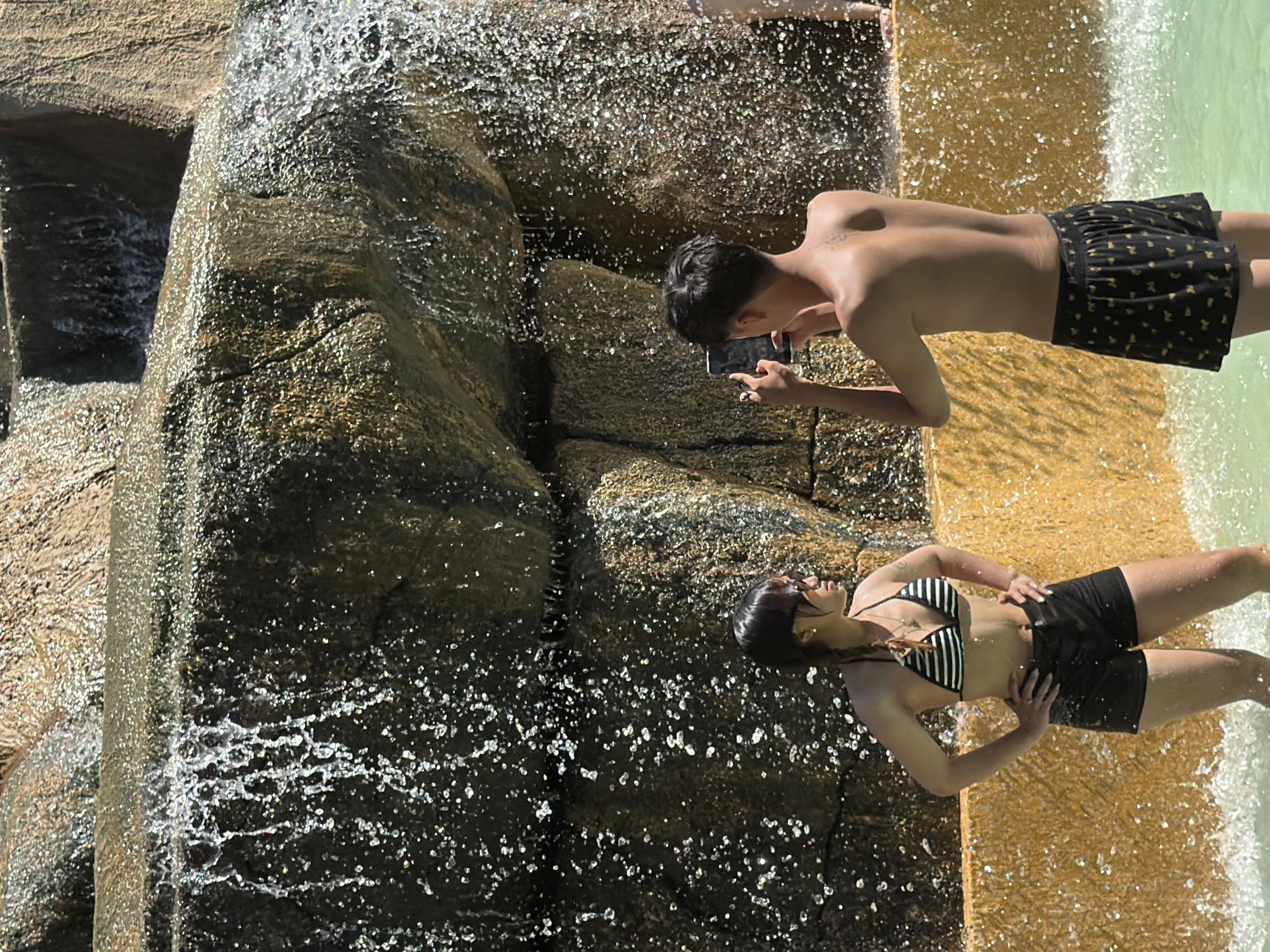 In Tay Nha Trang Ward, Vietnam, a young man and woman are enjoying a sunny day near a waterfall. The man, with short, dark hair, is shirtless and wearing black shorts with a gold pattern. He is holding a smartphone and appears to be taking a photo or video. The woman is wearing a black and white striped bikini and black shorts. Her hair is dark and braided. She has her hands on her hips and looks towards the camera.
The scene is a rocky waterfall with water cascading down. The rocks are wet and glistening, reflecting the sunlight. The water appears clear, and there are splashes and droplets visible. The ground around the waterfall is a sandy, yellowish color, also wet in places. The overall impression is one of a bright, warm, outdoor environment, likely a tourist attraction or a natural setting where people can cool off. The people's postures suggest they are relaxed and enjoying their time. The presence of a smartphone indicates modern technology is being used to capture the moment.