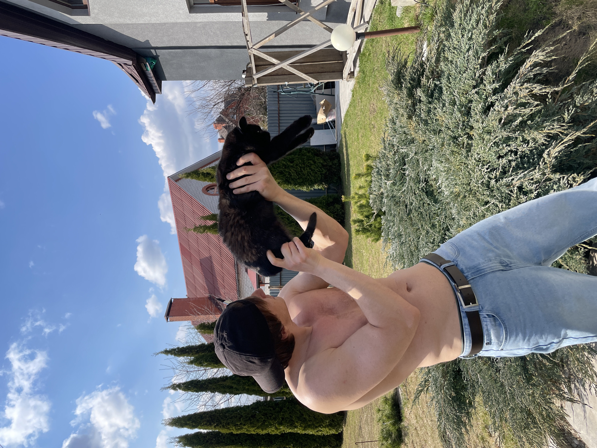 A shirtless man wearing jeans and a baseball cap is holding a black cat in his hands outdoors. The background shows a house with a red roof, a light post, trees, bushes, and a blue sky with clouds. There is no verifiable evidence to suggest the location is Kursk, Russia.